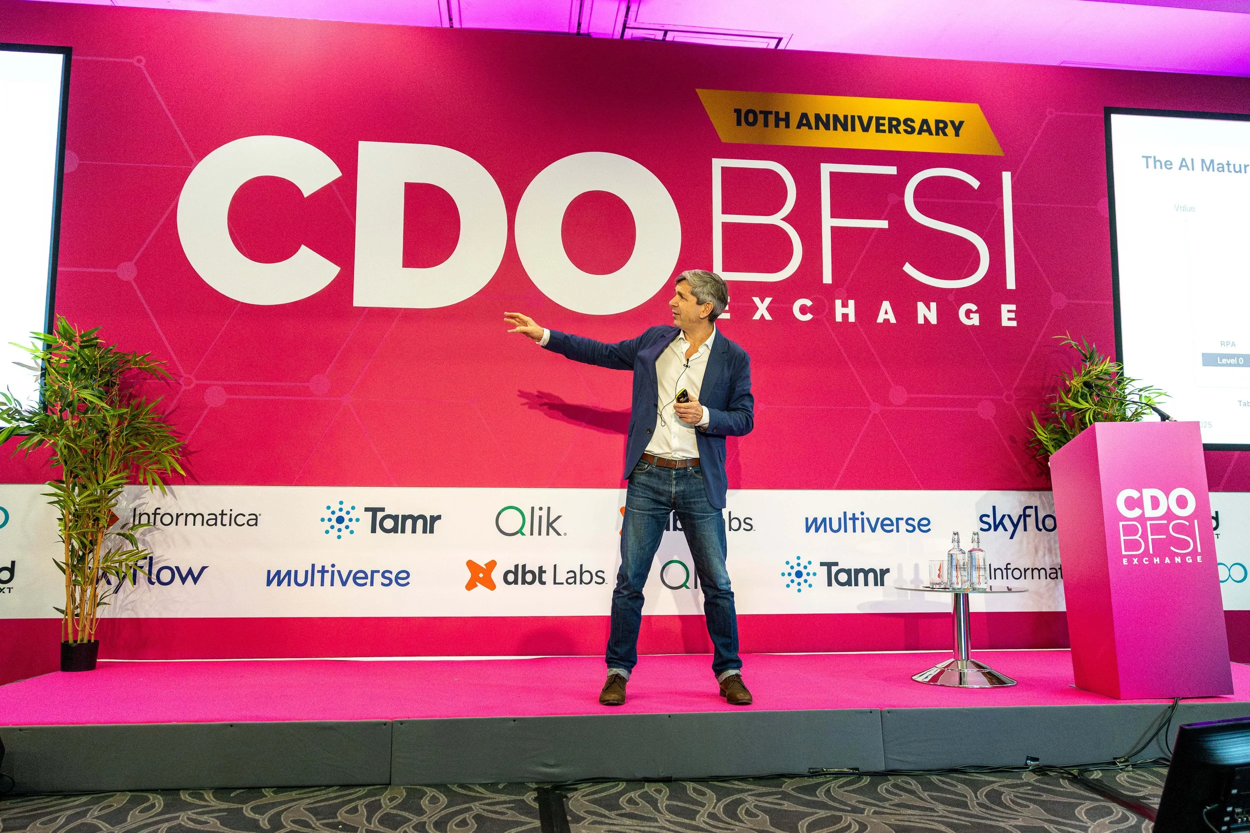 A man in a blue blazer and jeans giving a presentation on stage at the CDO BESI Exchange event, against a large pink backdrop with the text '10th Anniversary CDO BESI Exchange.' There are two screens on either side of the stage displaying presentatio
