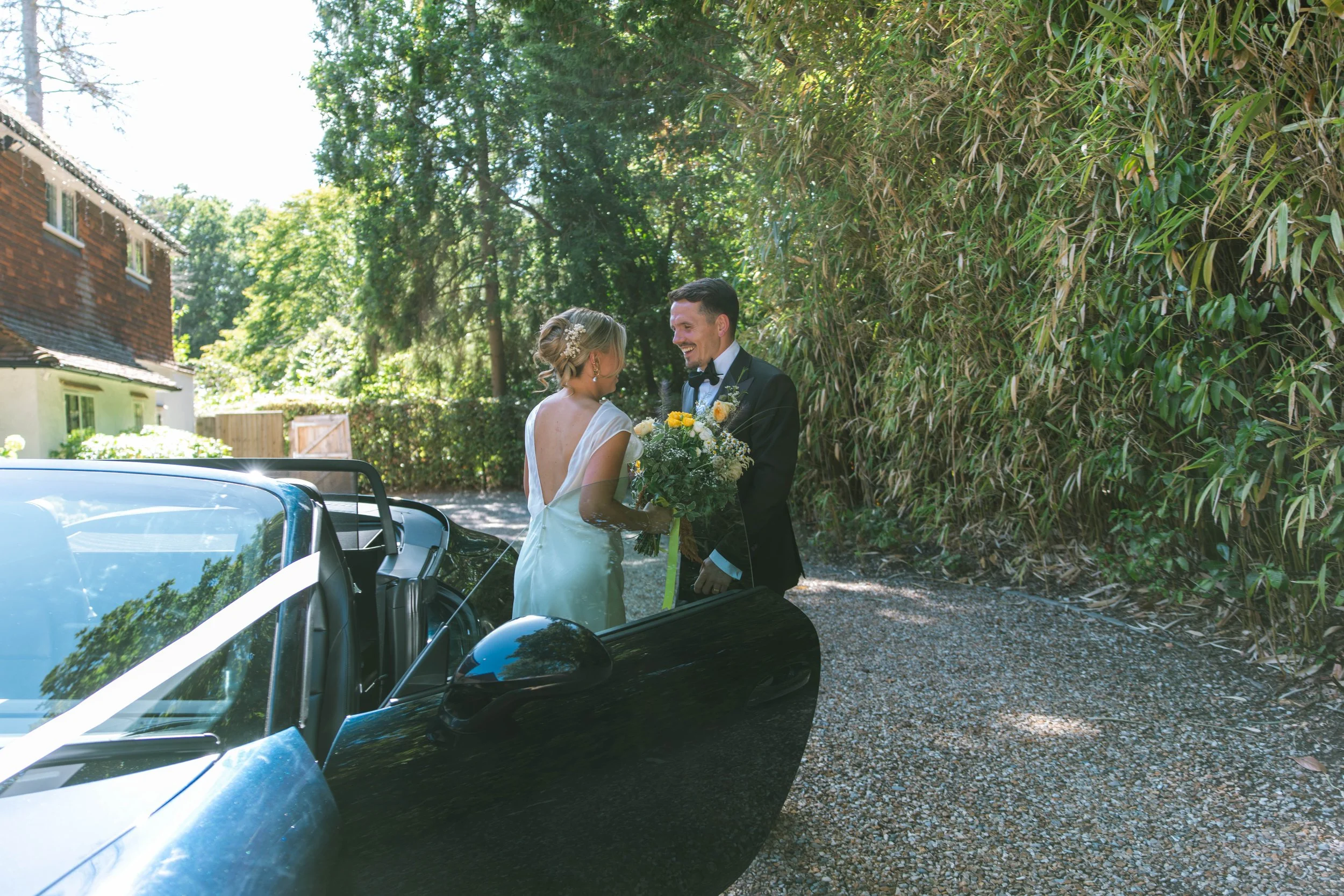 A bride and groom smiling and talking outside near a black car, with lush green trees and a wooden fence in the background.