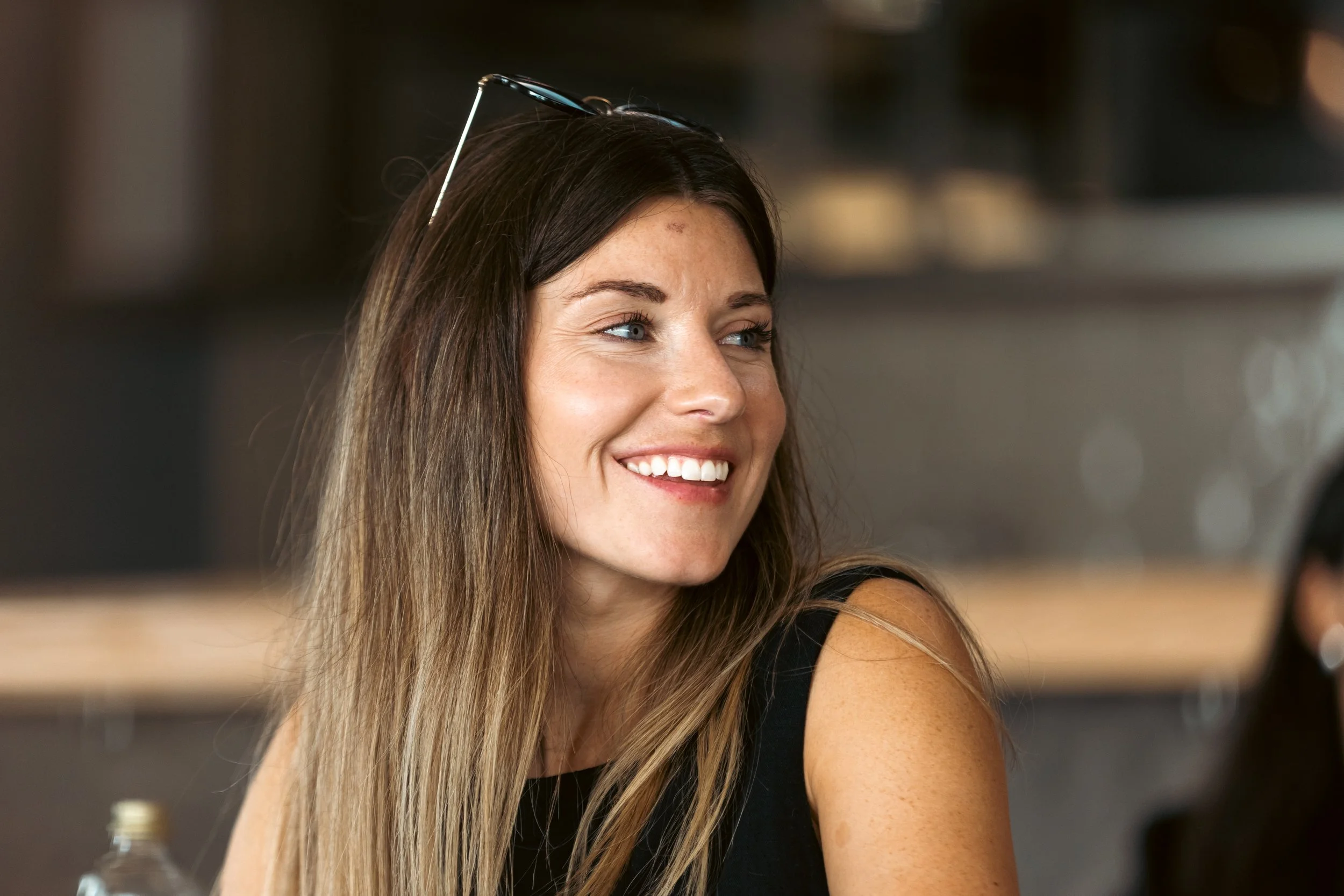 A woman with long brown hair and blue eyes smiling, wearing a black sleeveless top, with sunglasses resting on her head.