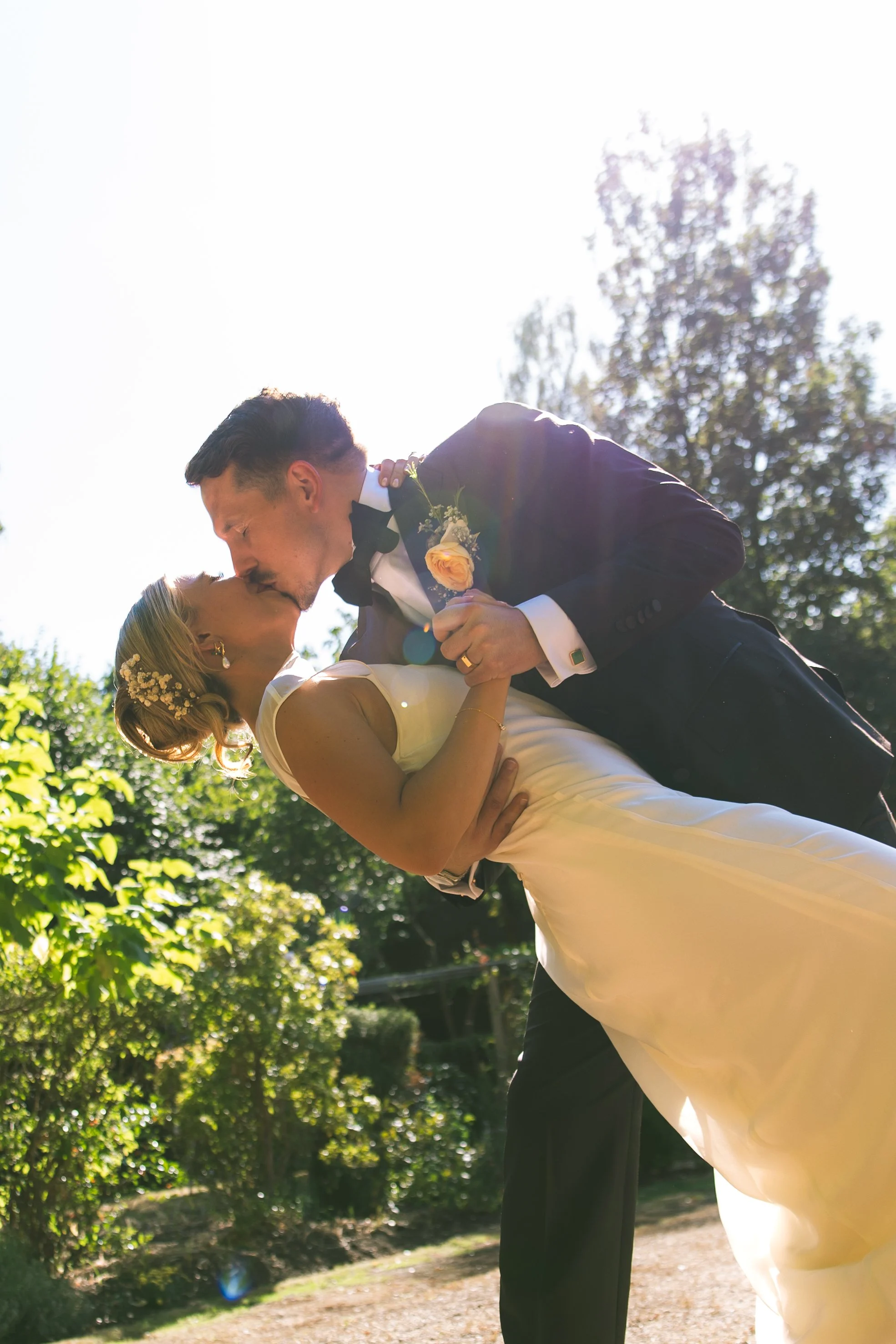 A groom dips his bride and kisses her during their wedding outdoor ceremony on a sunny day with green trees in the background.