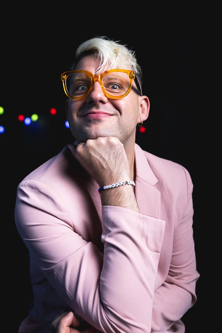 A person with blond hair, wearing oversized yellow glasses, and a pink jacket, smiling at the camera with a dark background with colorful blurred lights.