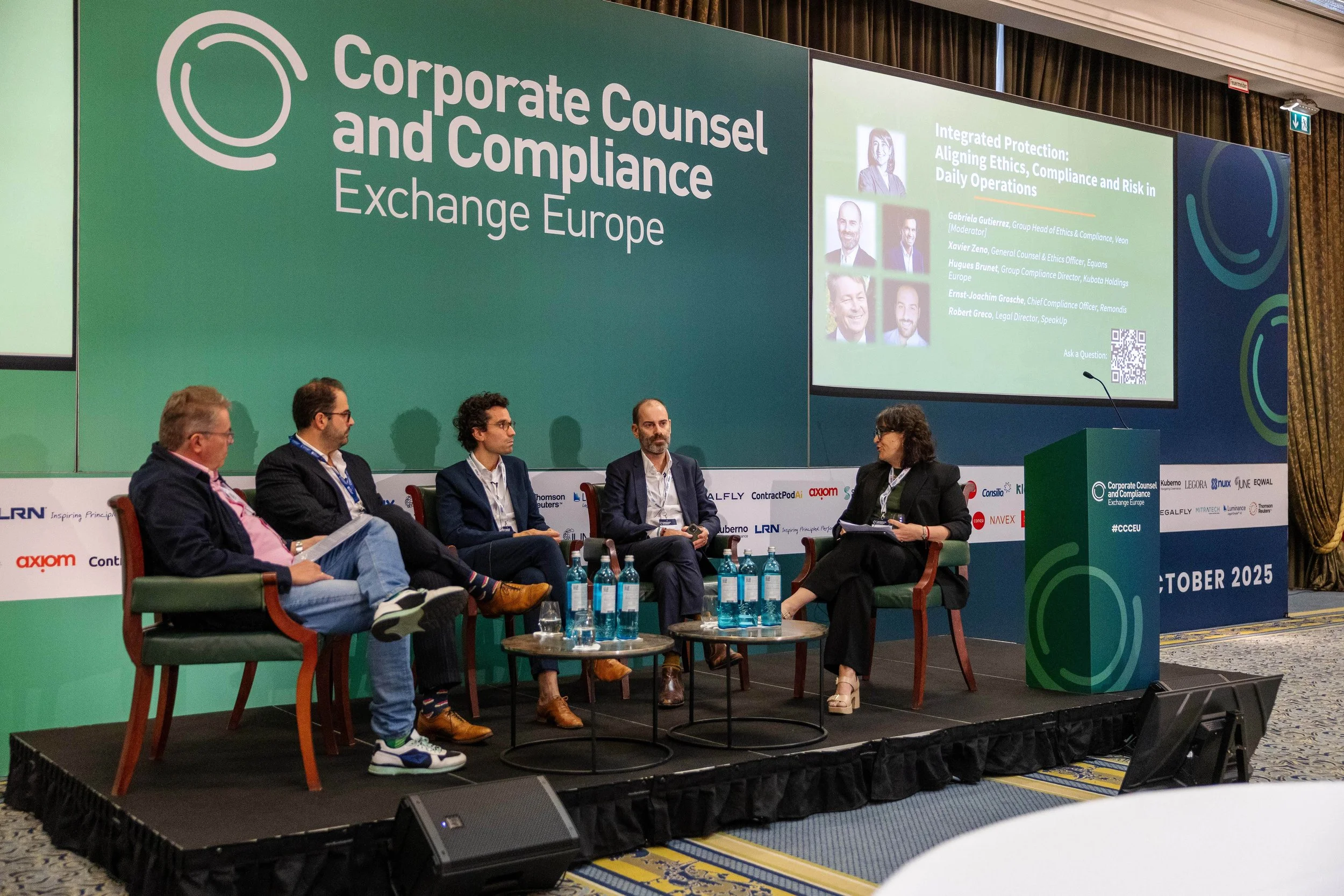 Panel discussion at a conference titled 'Corporate Counsel and Compliance Exchange Europe'. Five people are seated on stage, with some holding documents and water bottles placed on small tables in front of them. A large screen displays panel informat