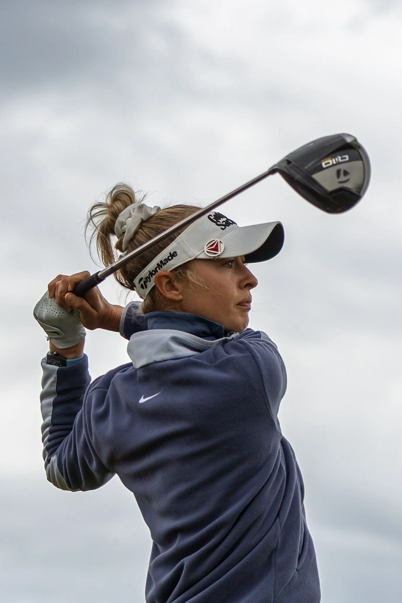 A woman playing golf, holding a golf club and about to swing, wearing a white visor and a navy and gray Nike jacket, with an overcast sky in the background.
