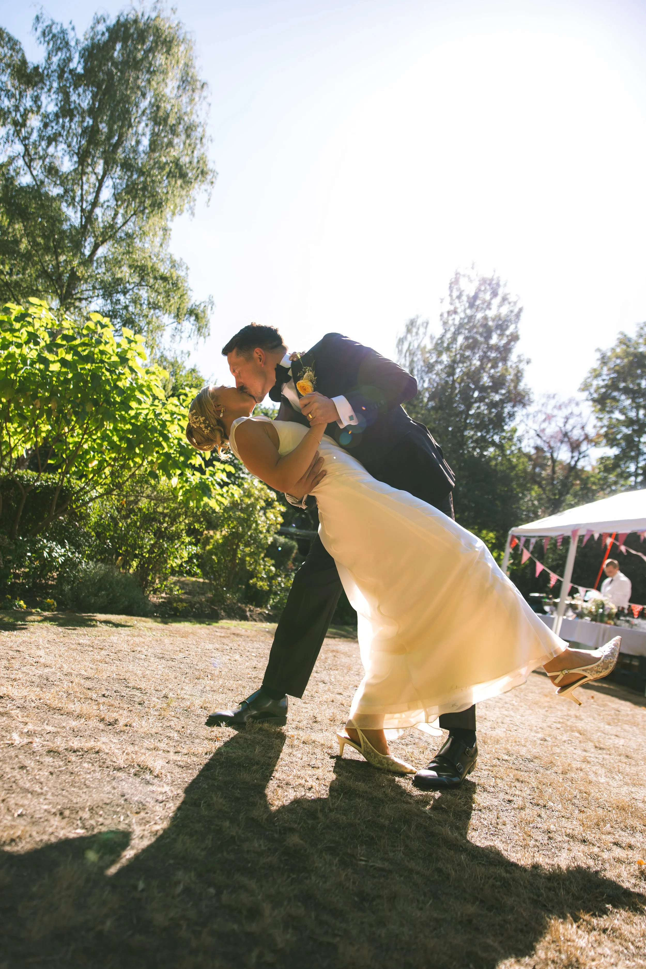 A bride and groom sharing a dance outdoors during daylight, with greenery and trees in the background. The groom is dipping the bride as they kiss, she is wearing a white wedding dress, he is in a dark tuxedo, and both are smiling. Shadows are cast o