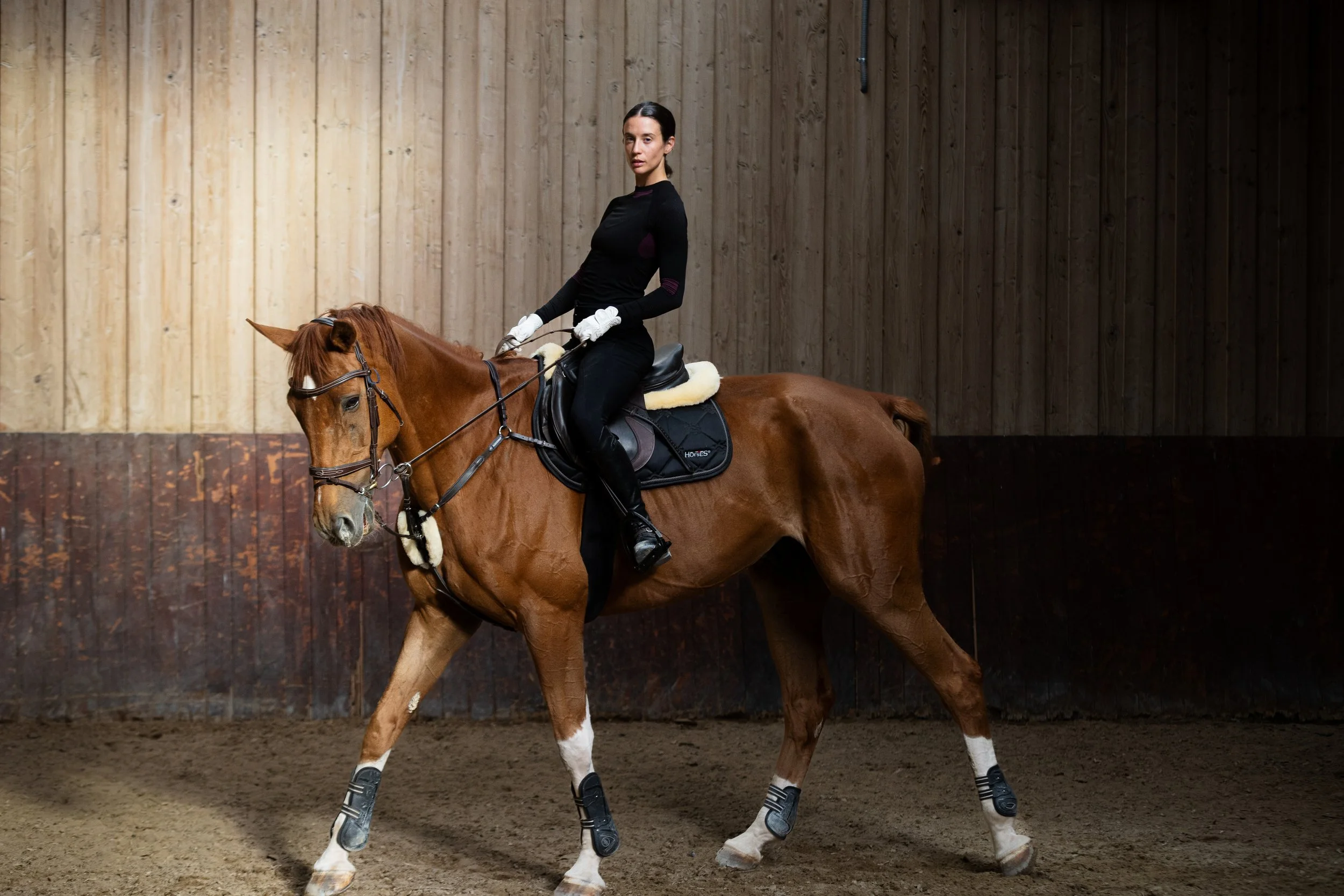 A woman in riding attire riding a brown horse inside an indoor riding arena with wooden walls.