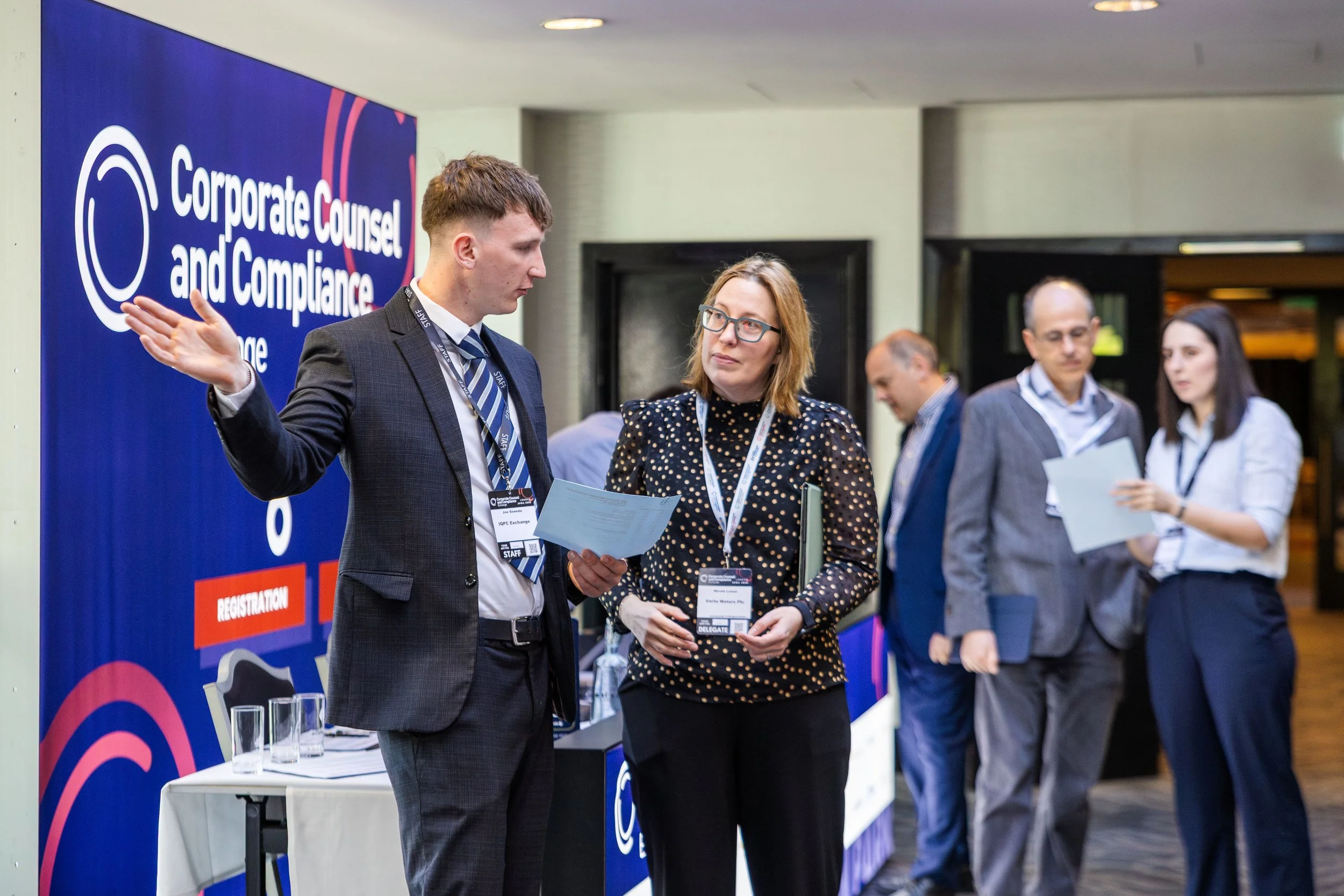 A man in a suit talking to a woman with glasses at a conference registration booth with a sign that reads 'Corporate Counsel and Compliance'. Four glasses of water are on the table.