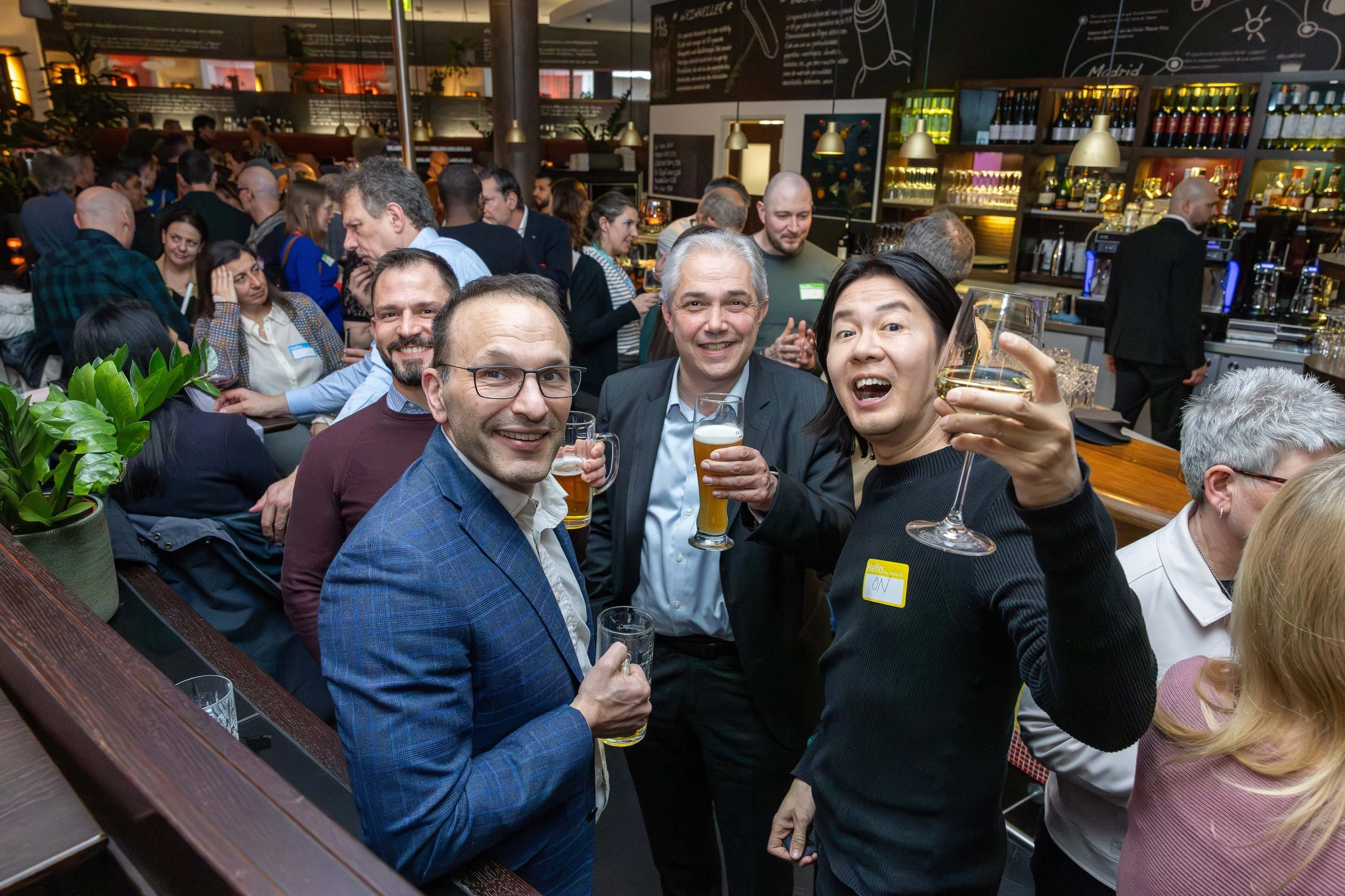 People at a lively indoor bar or restaurant, smiling, holding drinks, and enjoying a social gathering.
