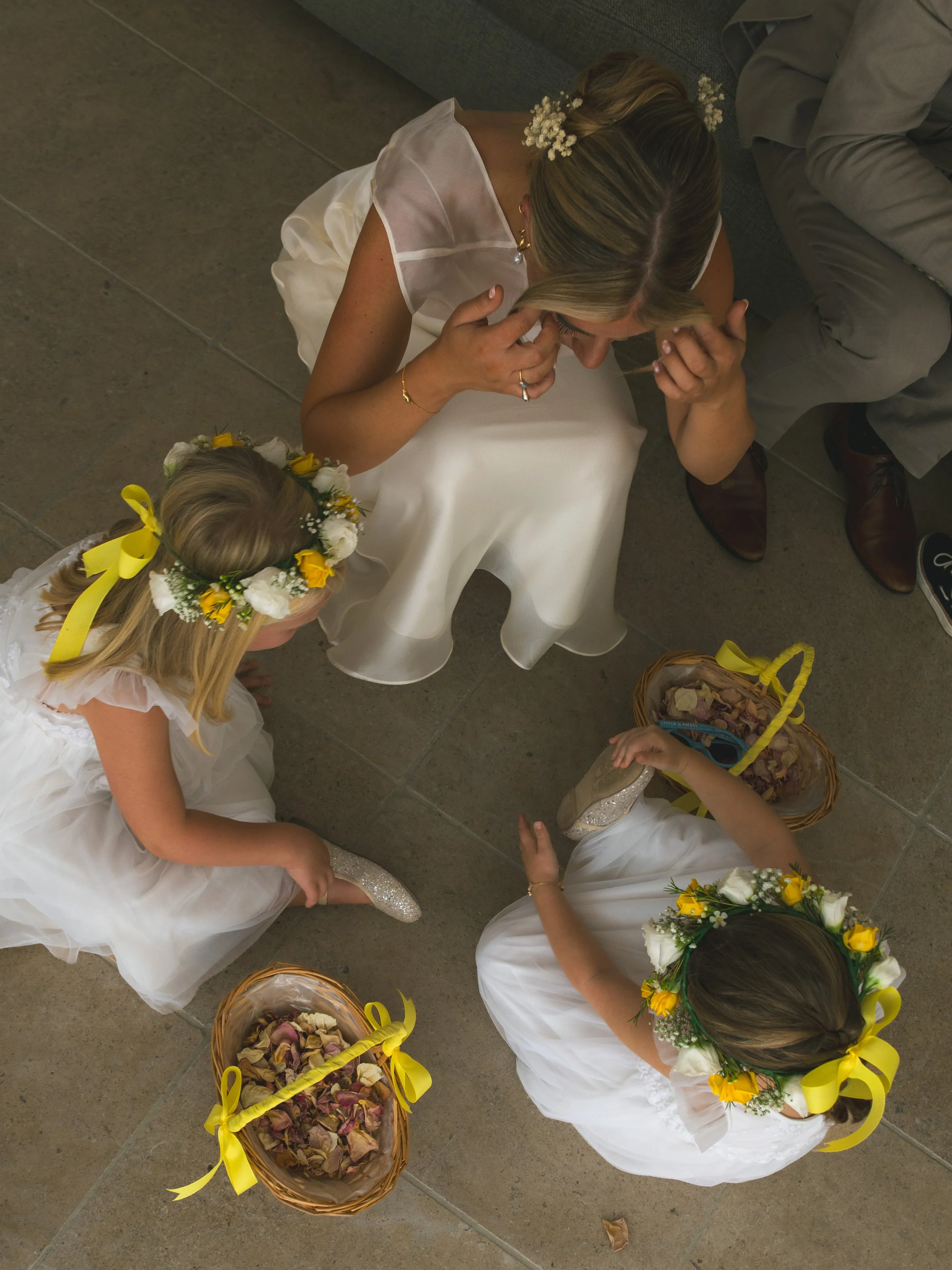 Children dressed in white, wearing flower crowns and bows, sitting on the floor, with baskets of dried flower petals around them, likely during a wedding or similar celebration.