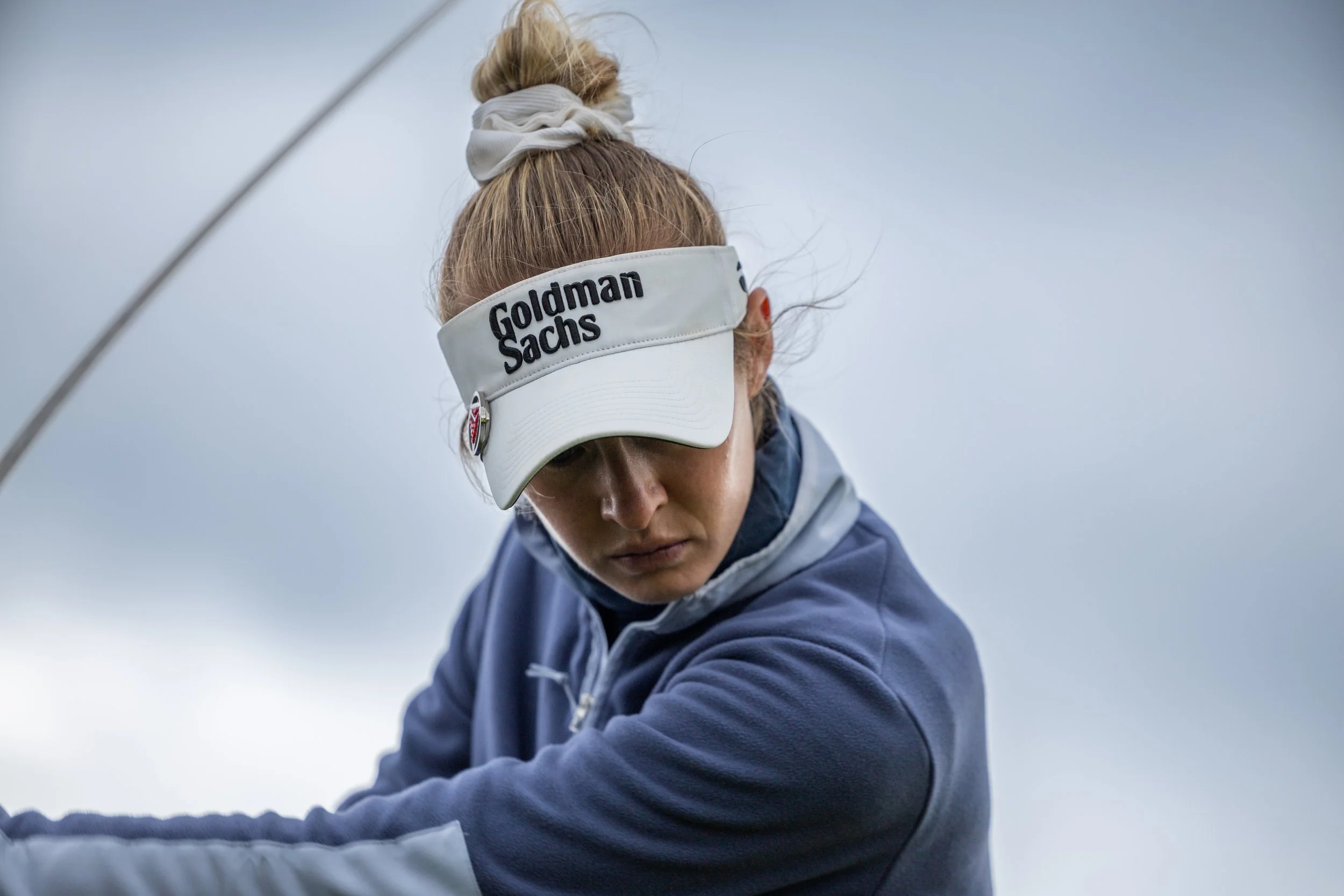 A woman wearing a Goldman Sachs visor cap and a blue jacket, focused on her activity outdoors on a cloudy day.