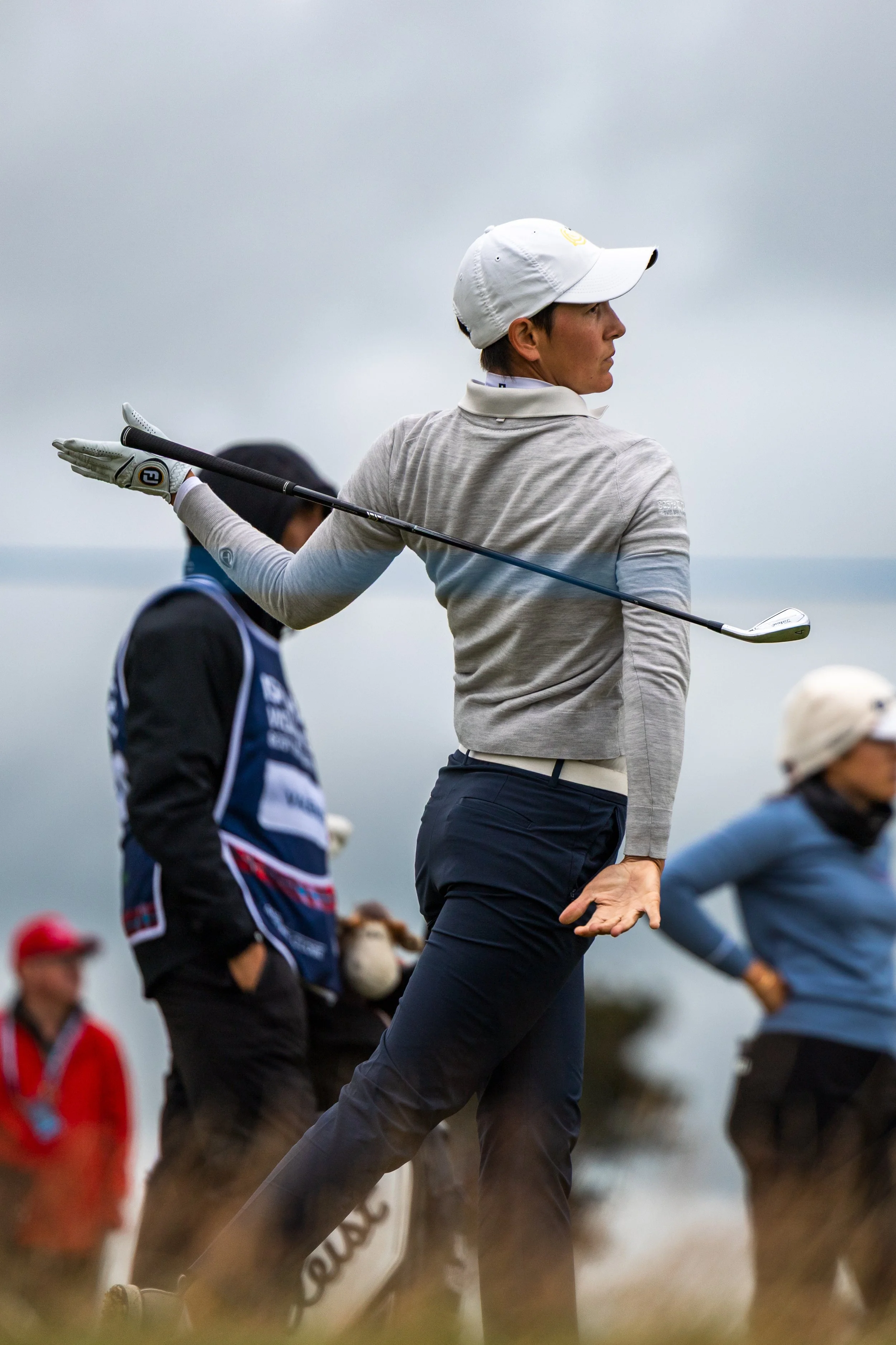 A woman playing golf on a cloudy day, holding a golf club, with other golfers and spectators in the background.