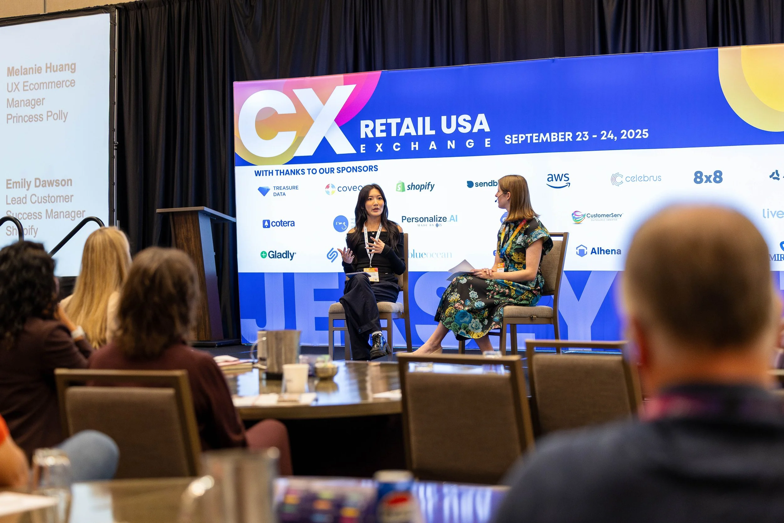 Two women sitting on stage during a conference or panel discussion at the Retail USA Exchange held on September 23-24, 2025. One woman with long dark hair is speaking, while the other with shoulder-length red hair listens. The large screen behind dis