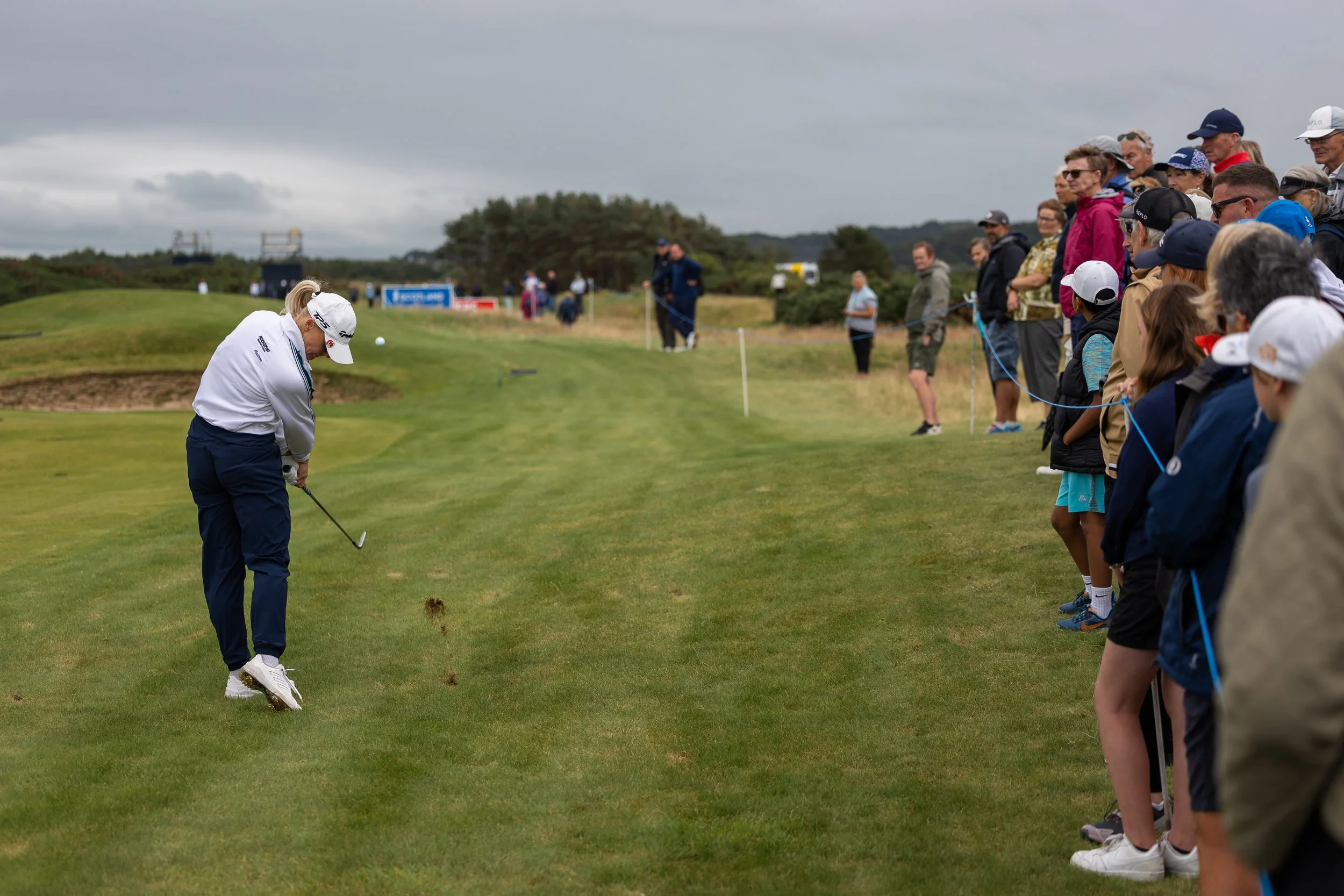 A female golfer in a white shirt and navy pants is putting on a golf course. She is wearing a white cap with a logo. A crowd of spectators, wearing casual clothing and caps, is watching her from the side of the green, standing behind a blue rope barr
