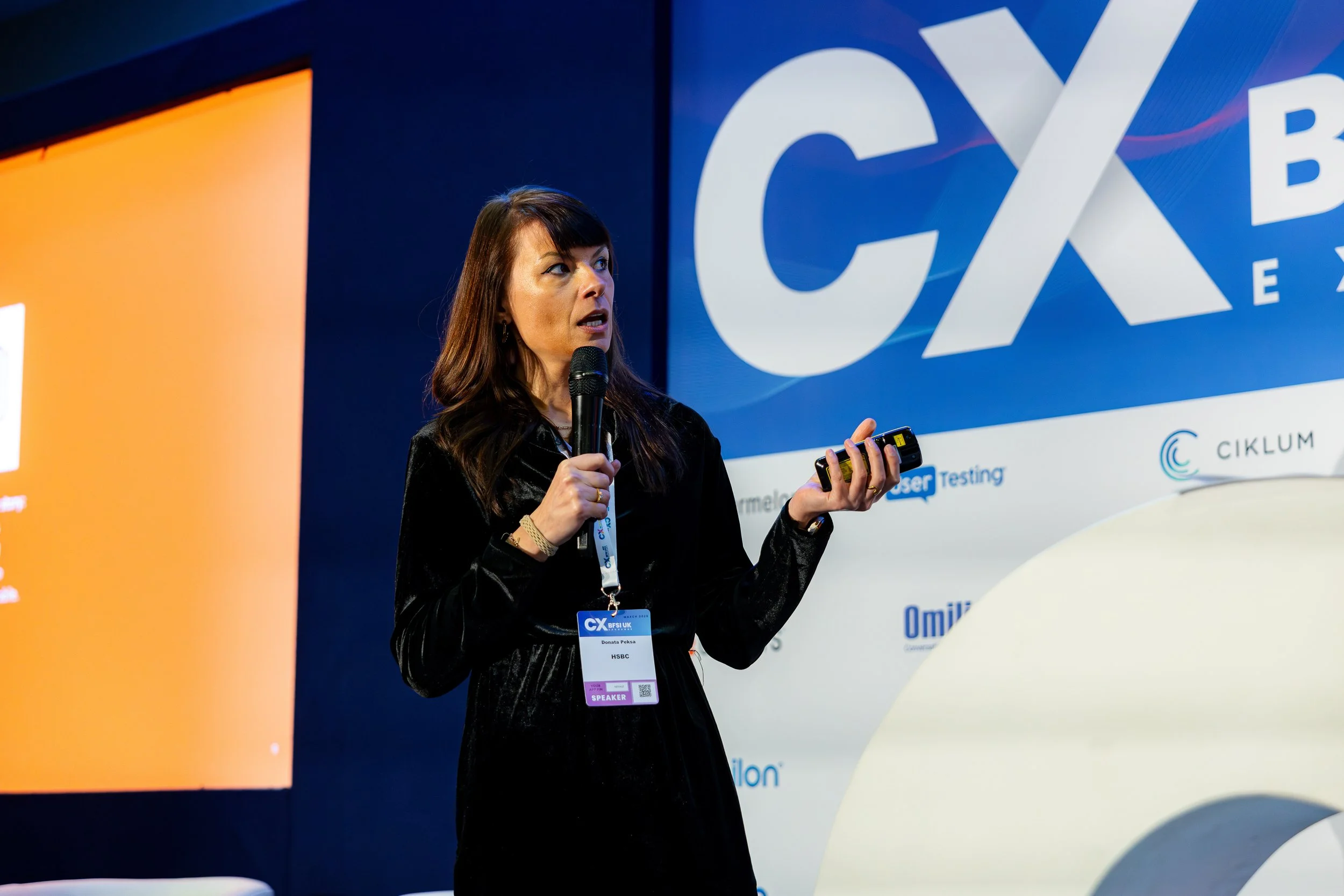 A woman with dark hair is giving a presentation or speech at a conference, standing in front of a large screen with a blue and white logo that reads 'CX' and other text. She is holding a microphone in her right hand and a small device, possibly a rem
