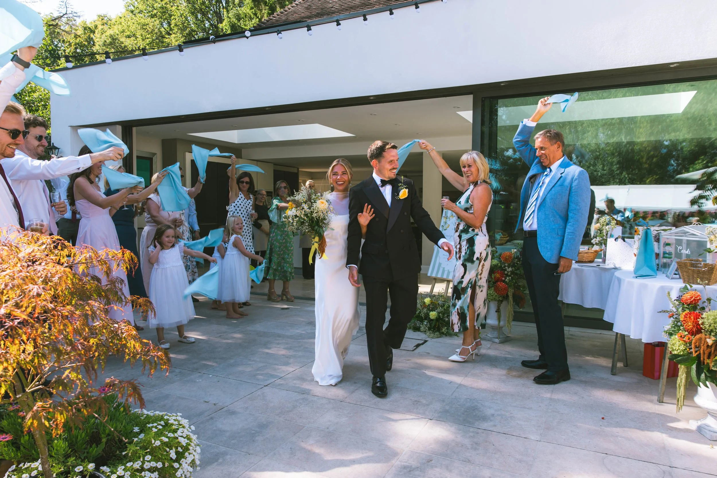 Wedding celebration outdoors with guests throwing blue cloth napkins in the air as a bride and groom walk hand in hand. The bride holds a bouquet of flowers, and the groom wears a suit with a boutonniere. Guests are smiling, dressed in semi-formal at