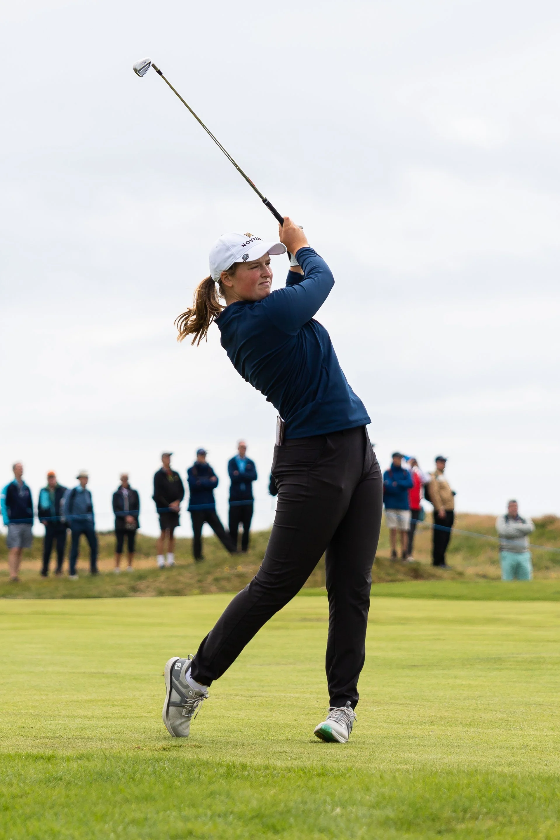 A female golfer in a navy blue long sleeve shirt and black pants swings a golf club on the green, with spectators in the background.