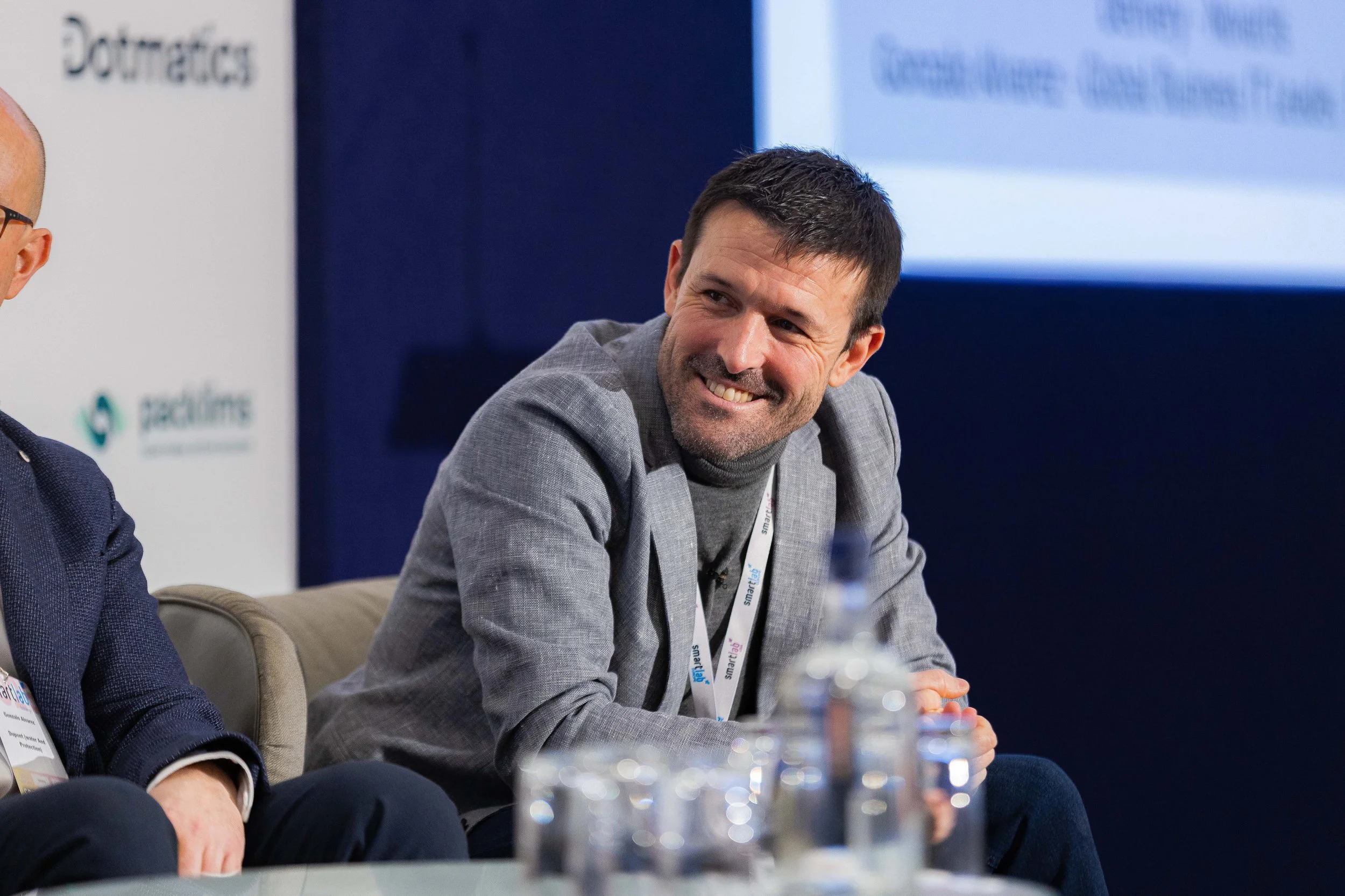 A man with short dark hair, a beard, and a gray blazer smiling while sitting at a panel discussion during a conference.