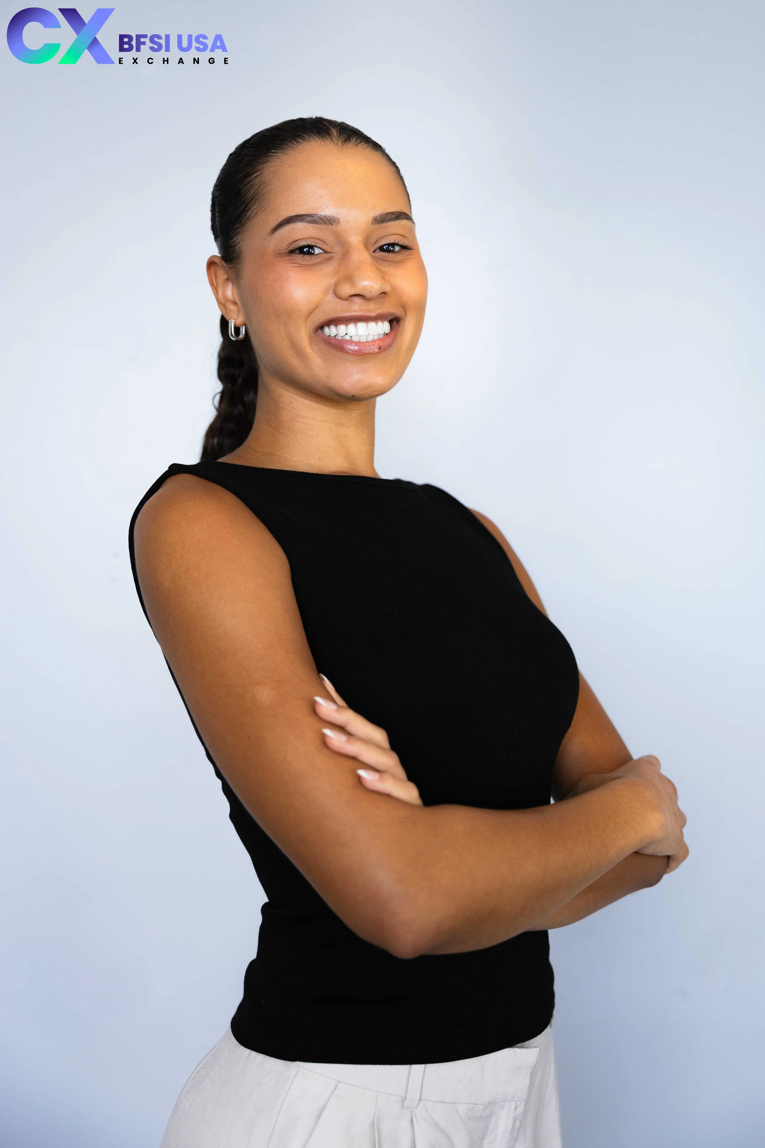 A woman smiling with arms crossed, wearing a black sleeveless top and light-colored pants, standing against a plain light blue background with the logo of BFSI USA Exchange in the top left corner.