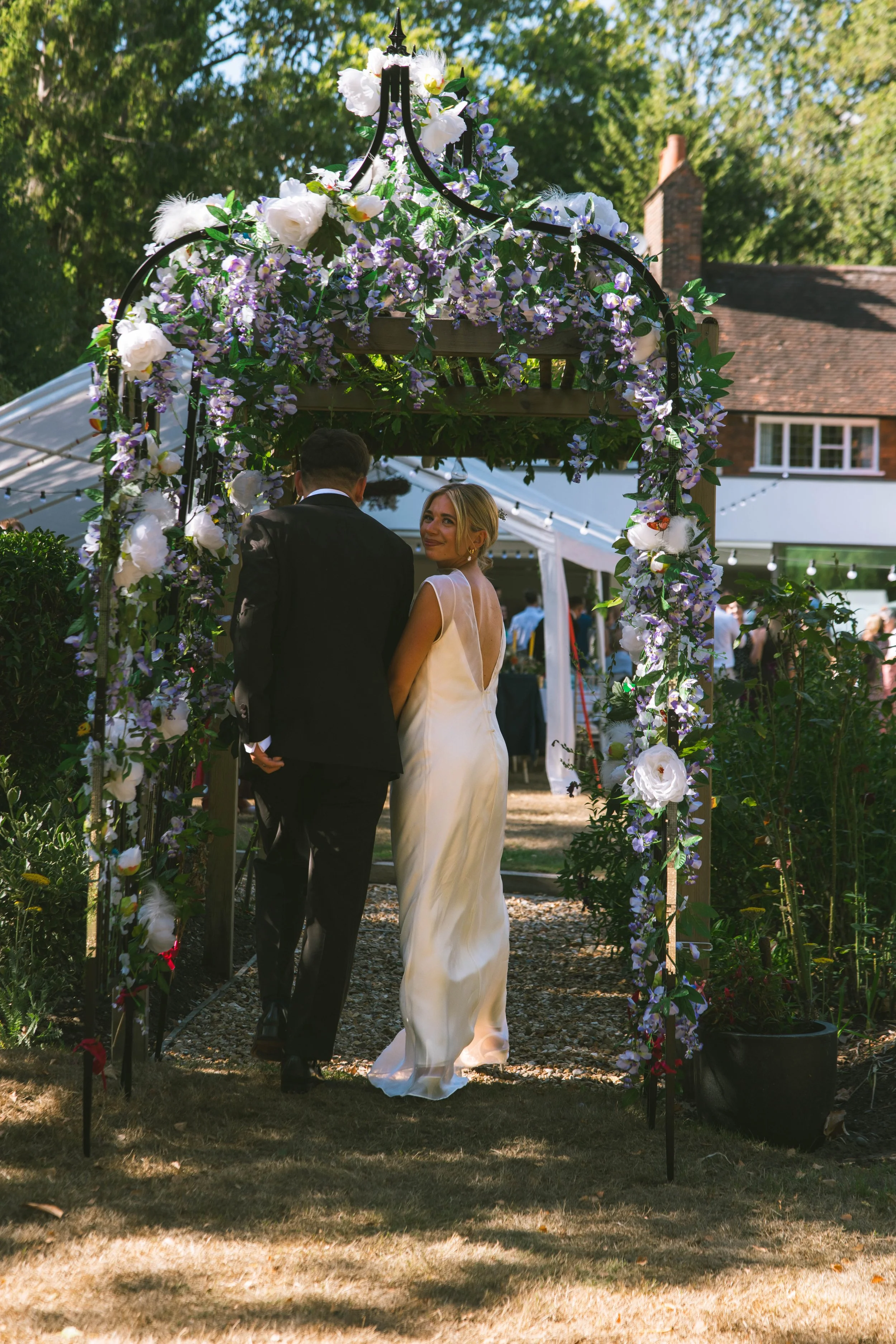 A bride and groom standing under a decorated floral wedding arch outdoors on a sunny day.