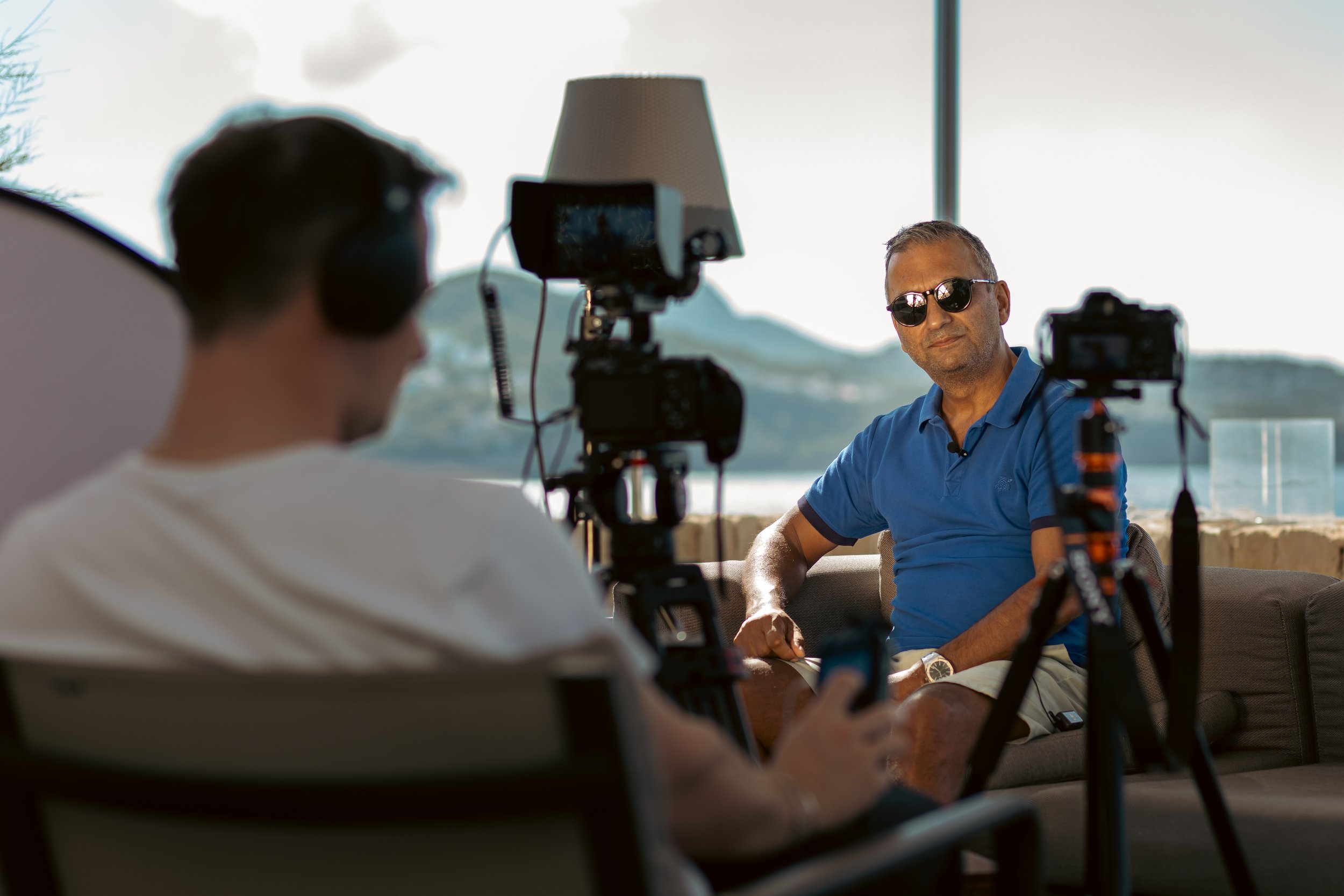 Two men are filming an interview outdoors, with one man wearing sunglasses and the other wearing headphones, sitting on a couch near a large window with a view of the outdoors.
