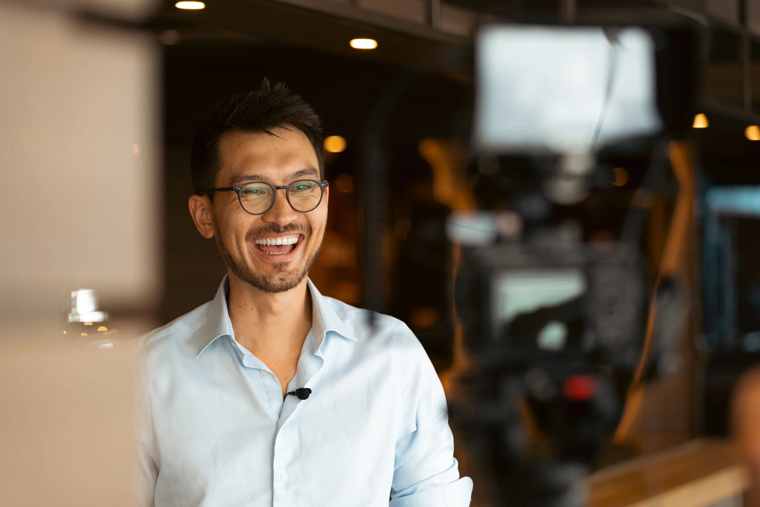 A smiling man with glasses and a light blue shirt standing in a modern office or tech environment with shelves and monitors in the background.