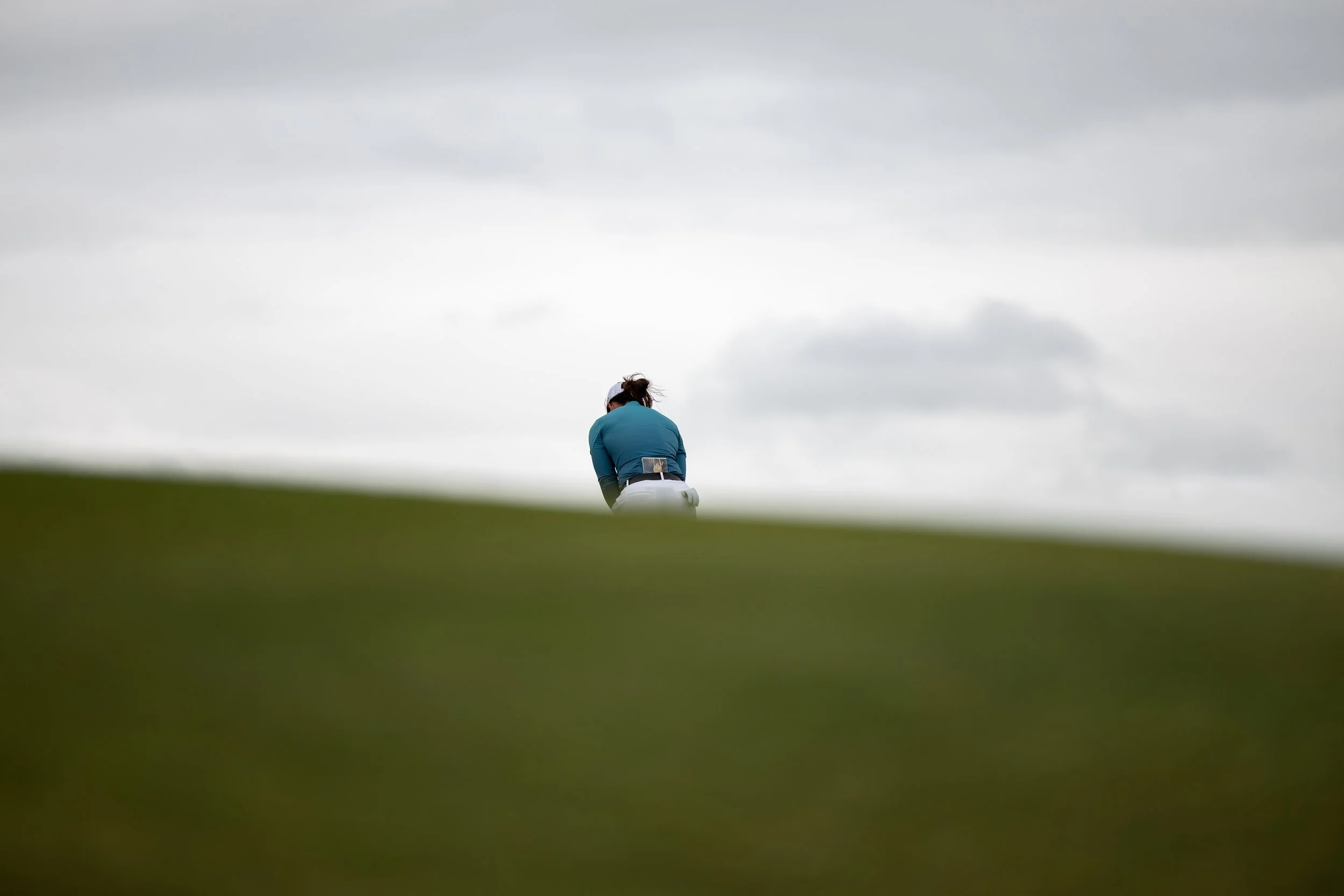 A golfer in a teal shirt and white cap bending over on a golf course, with a cloudy sky overhead and a blurred green foreground.