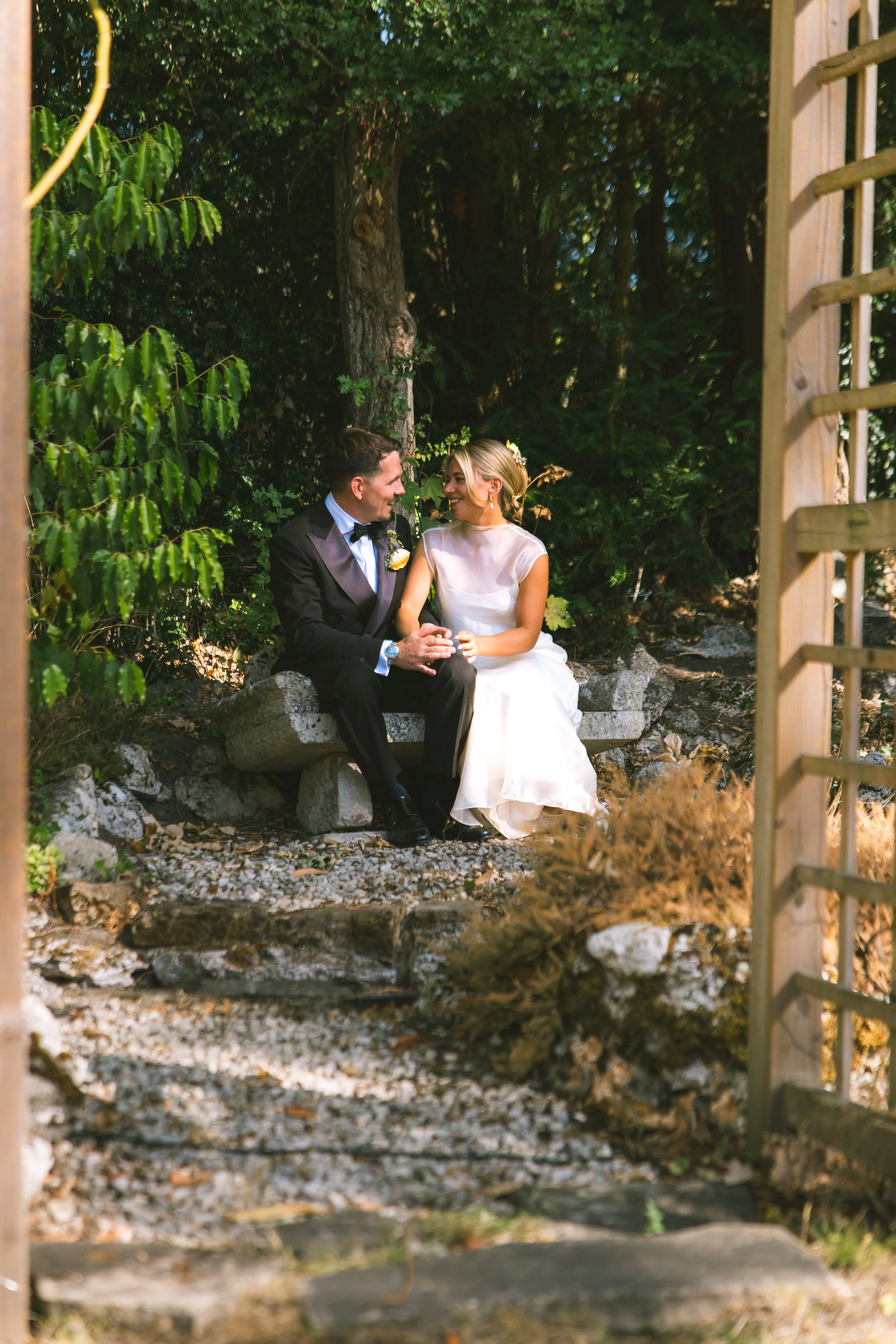 A bride and groom sitting on a stone bench in a wooded outdoor setting, smiling and holding hands, with trees and greenery around them.