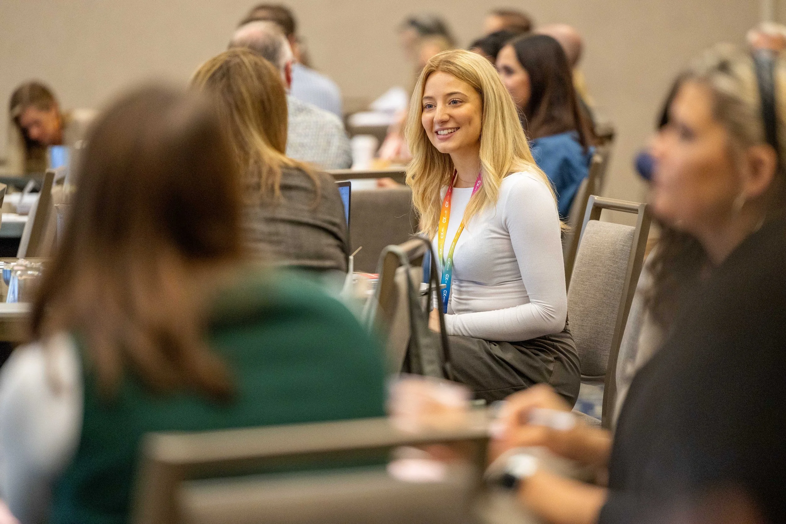 A young woman with blonde hair smiling and talking to a person across from her at a conference. She is wearing a white top and a colorful lanyard, and there are other people in the background.