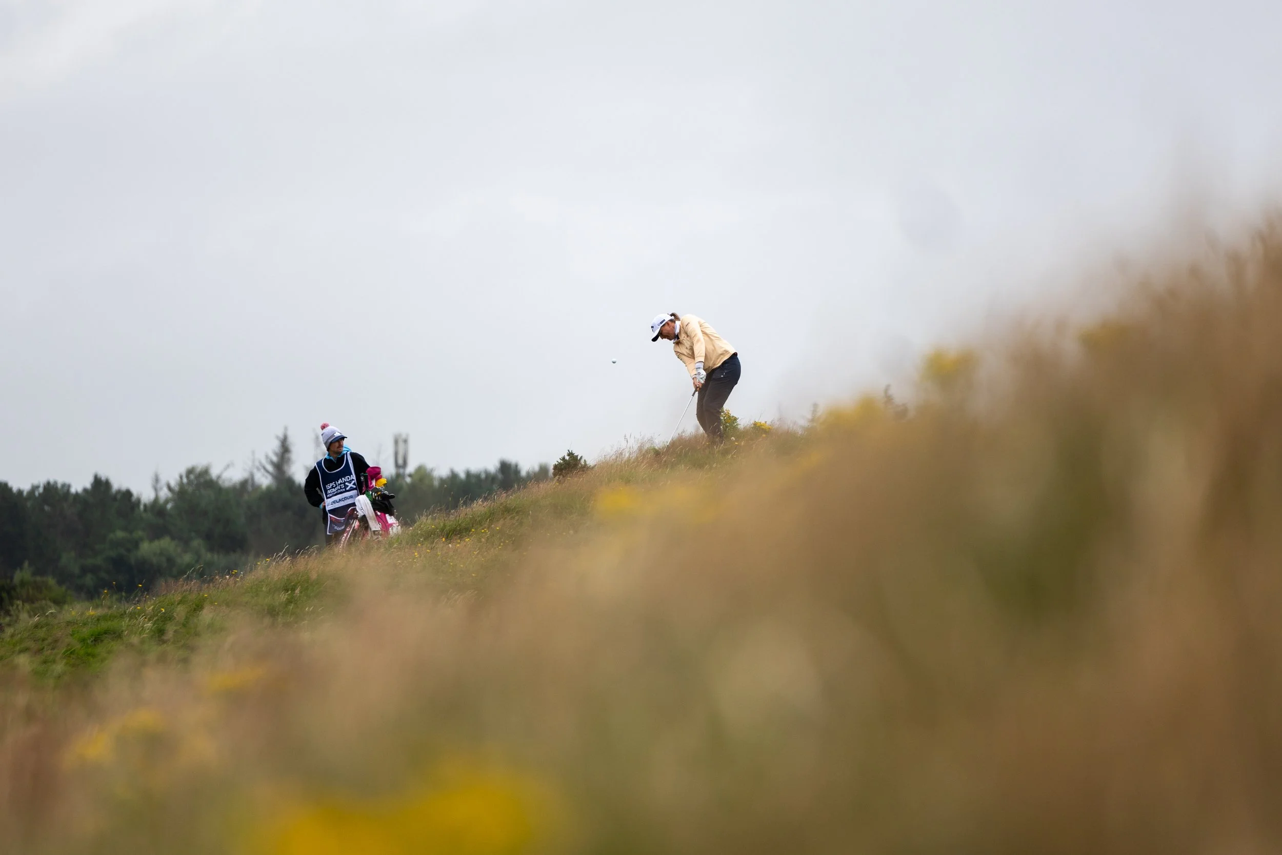 A golfer on a grassy golf course preparing to hit a ball, with a caddy nearby carrying golf clubs, set against a cloudy sky.