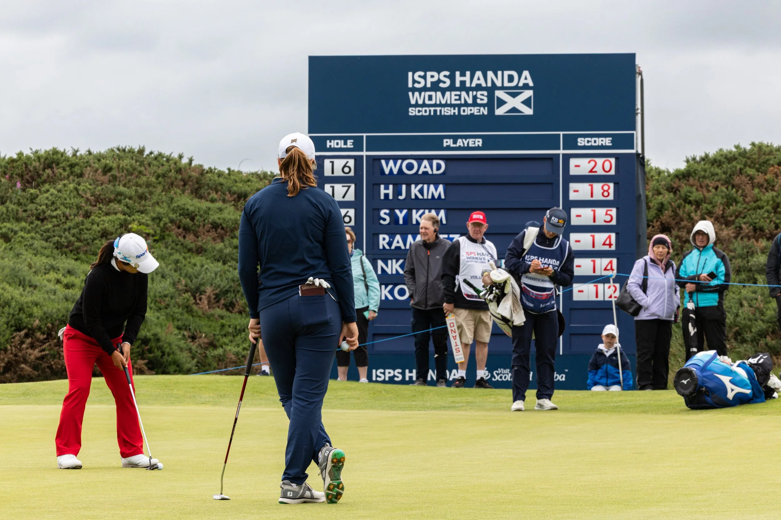 Women golfers on a green during the ISPS Handa Women's Scottish Open, with a scoreboard in the background and spectators watching.