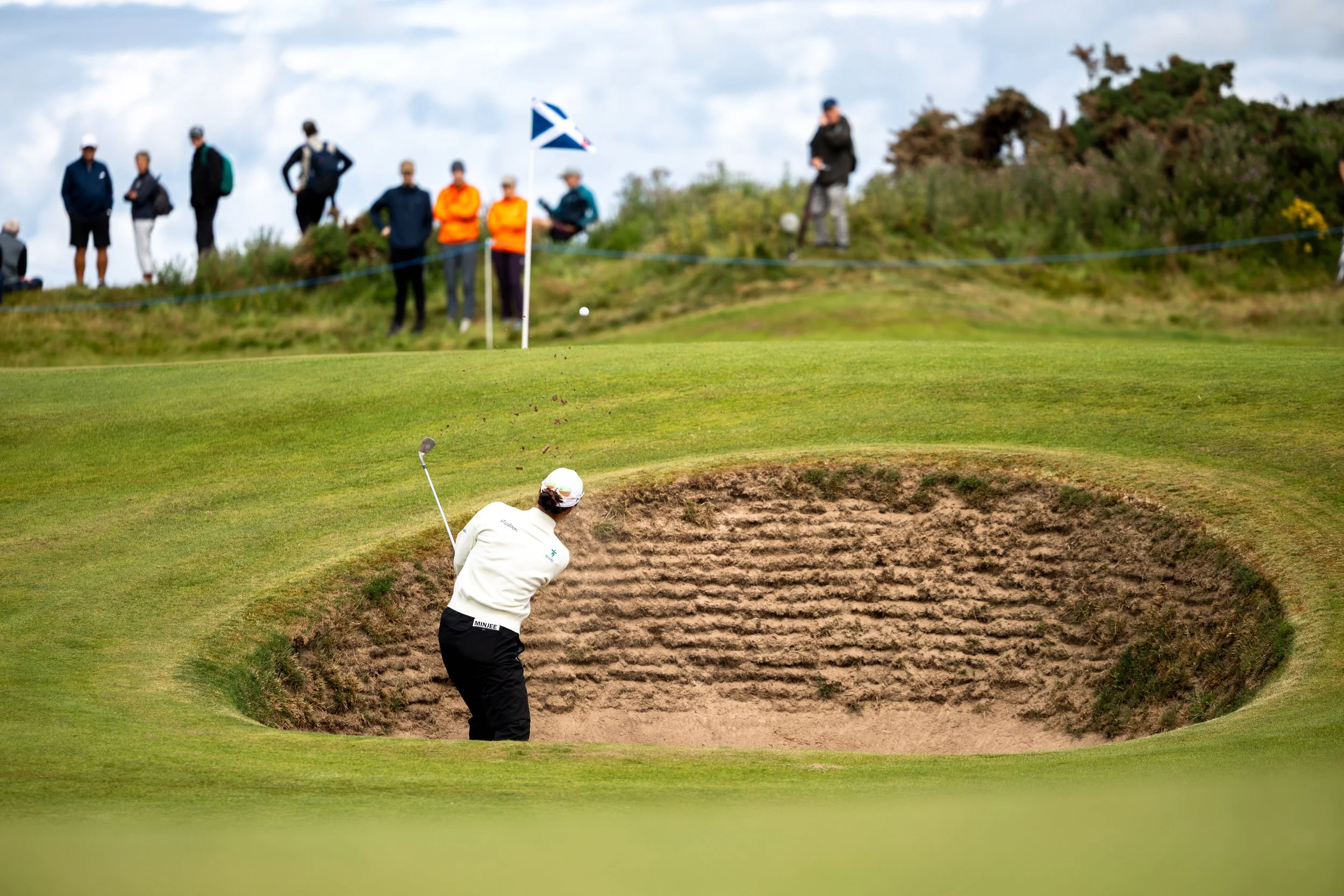 A golfer is taking a shot from a sand bunker on a golf course, while a group of spectators watch nearby.