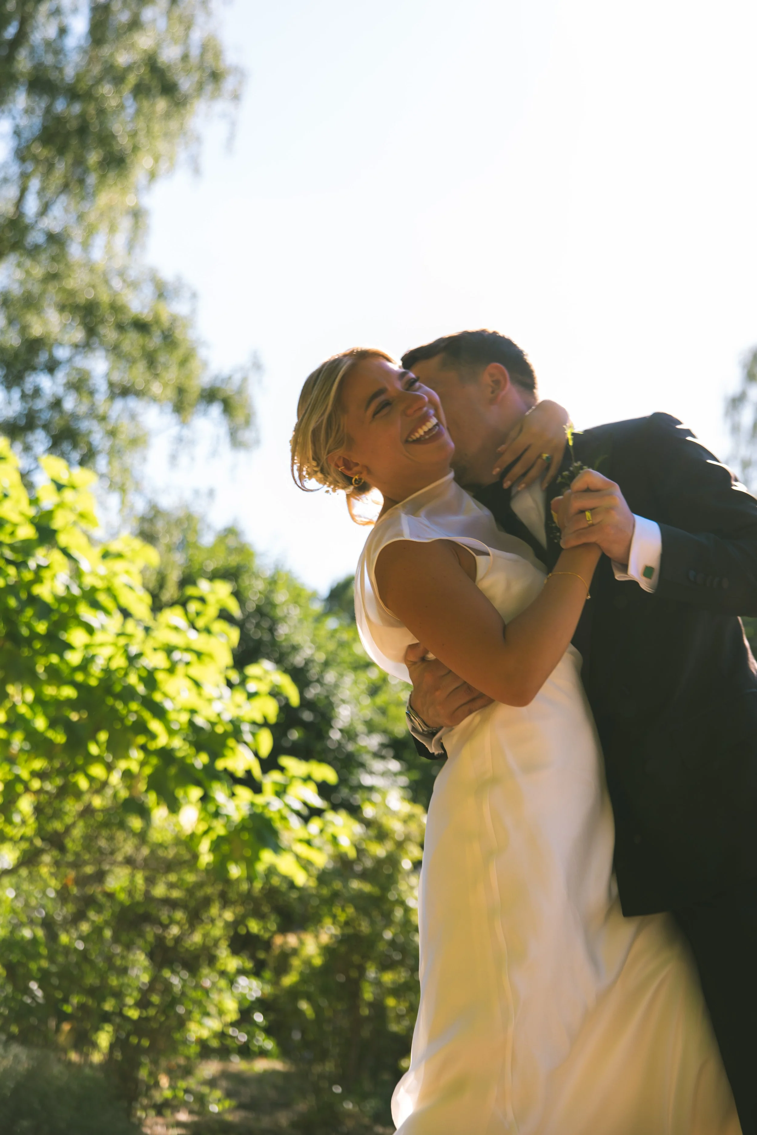 A couple, the woman in a white dress and the man in a dark suit, embracing and smiling outdoors with trees and bright sunlight in the background.