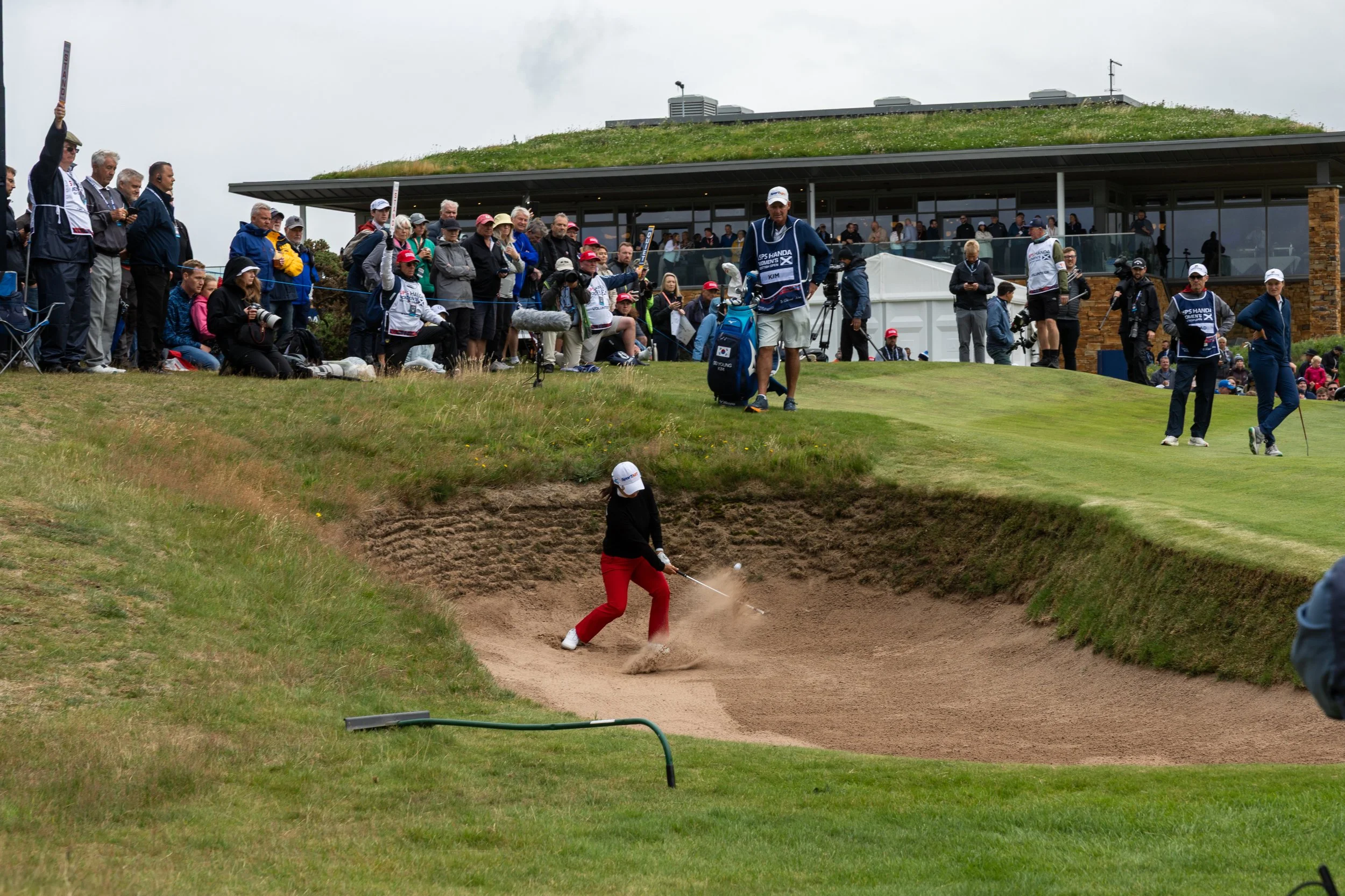 A woman in red pants and black top is hitting a golf ball from a sand bunker on a golf course, with focused spectators and photographers watching nearby.