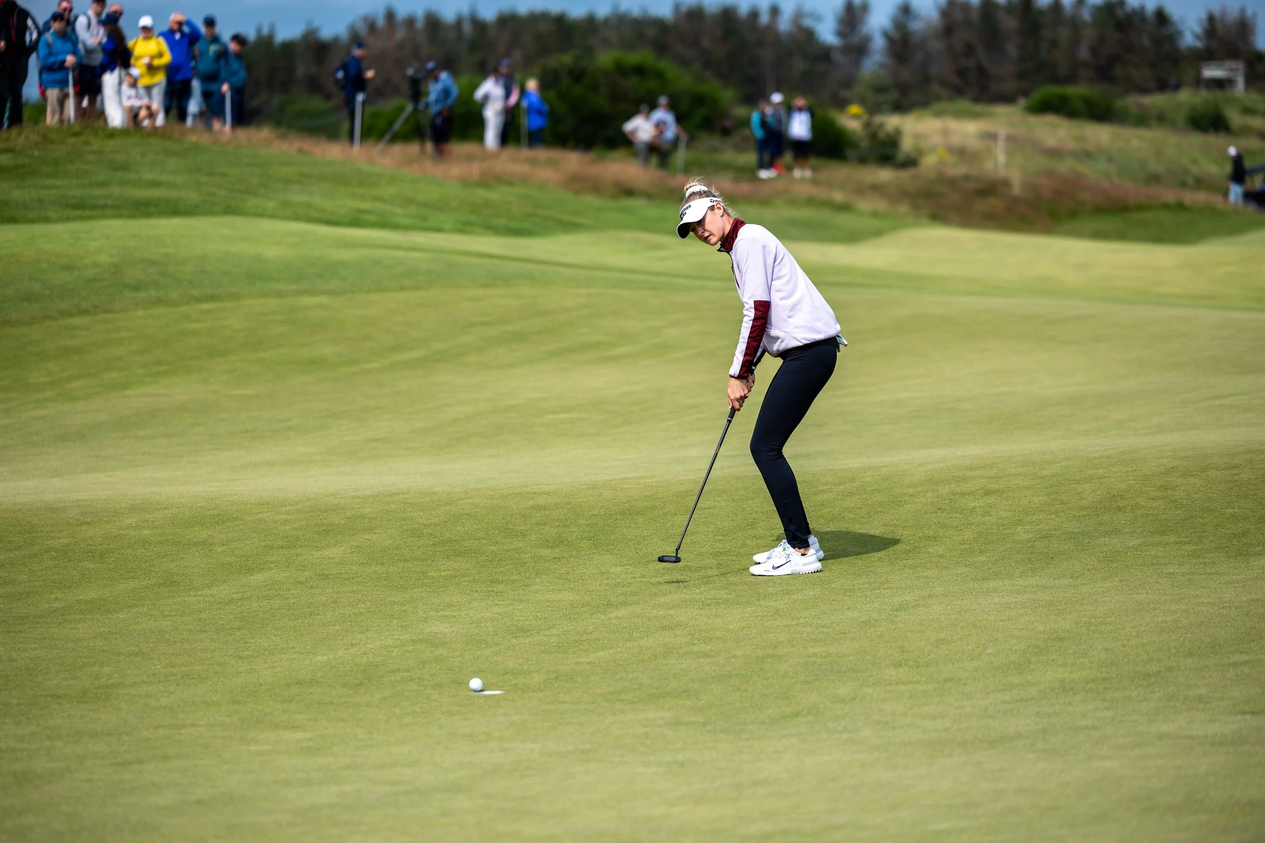 A female golfer in a white jacket with maroon sleeves taking a shot on a golf course while a group of spectators watch from a distance.