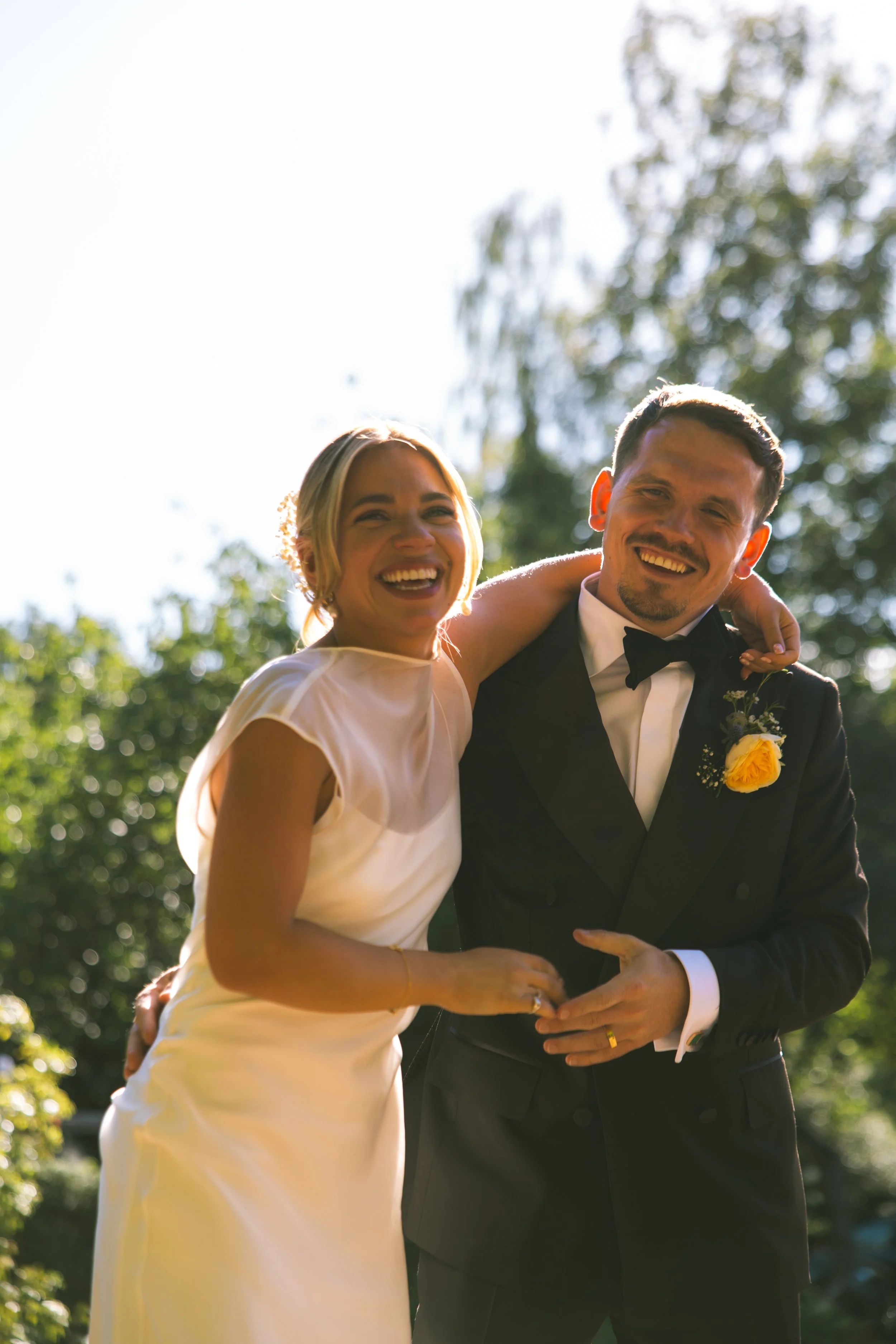 A smiling bride and groom dressed in wedding attire, standing outdoors under a clear sky with trees in the background. The bride wears a white dress and the groom wears a tuxedo with a yellow boutonniere.