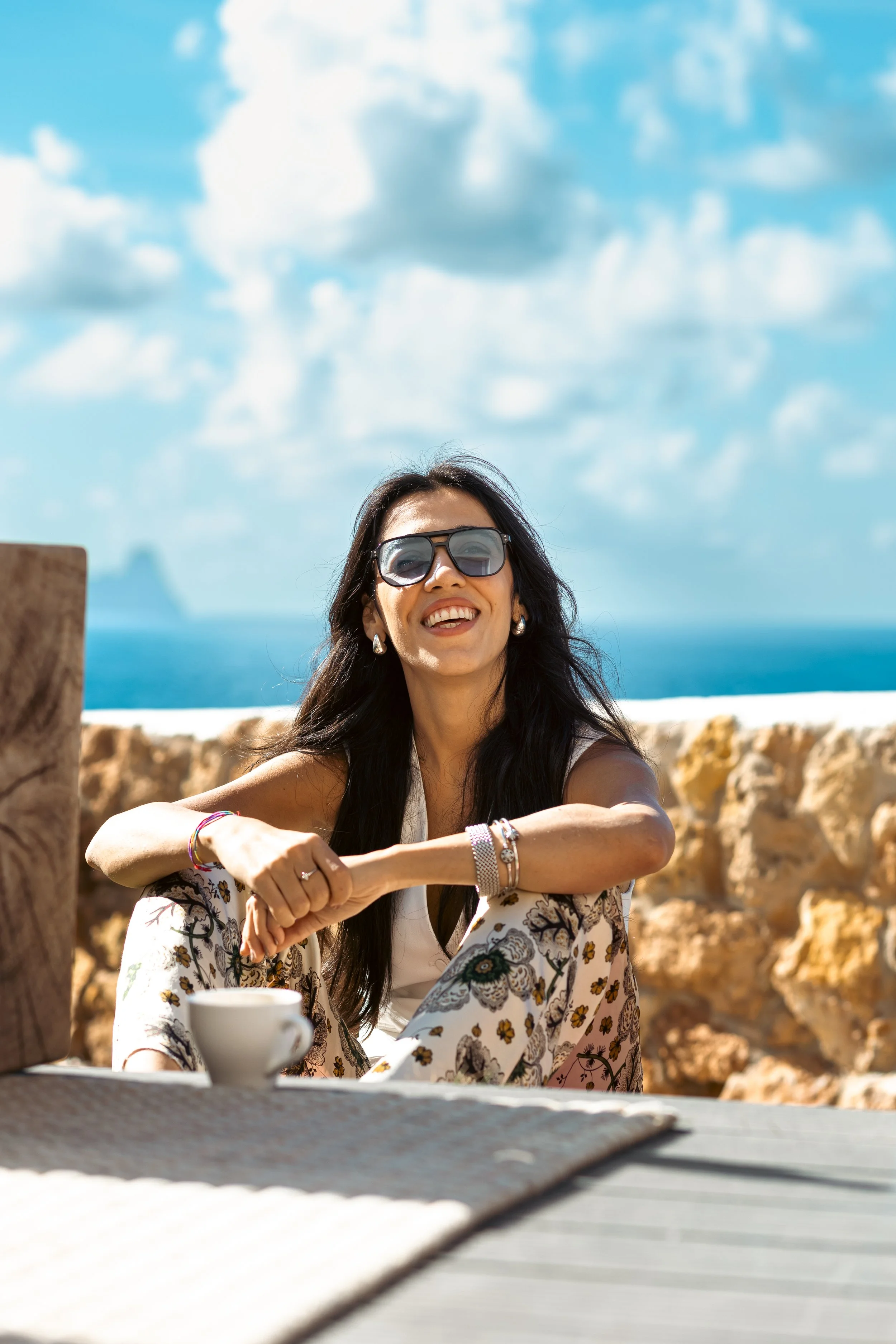A woman with long dark hair, wearing sunglasses, a white top, and a floral kimono, smiling while sitting outdoors at a table with a teacup, against a backdrop of rocks, blue sky, and the ocean.