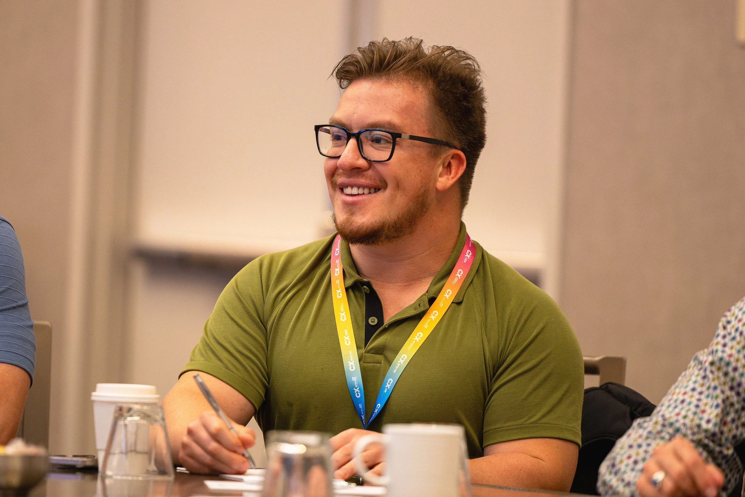 A man with glasses and a beard smiling, wearing a green polo shirt and a yellow and blue lanyard, seated at a table during a meeting or conference, with cups and a paper on the table.
