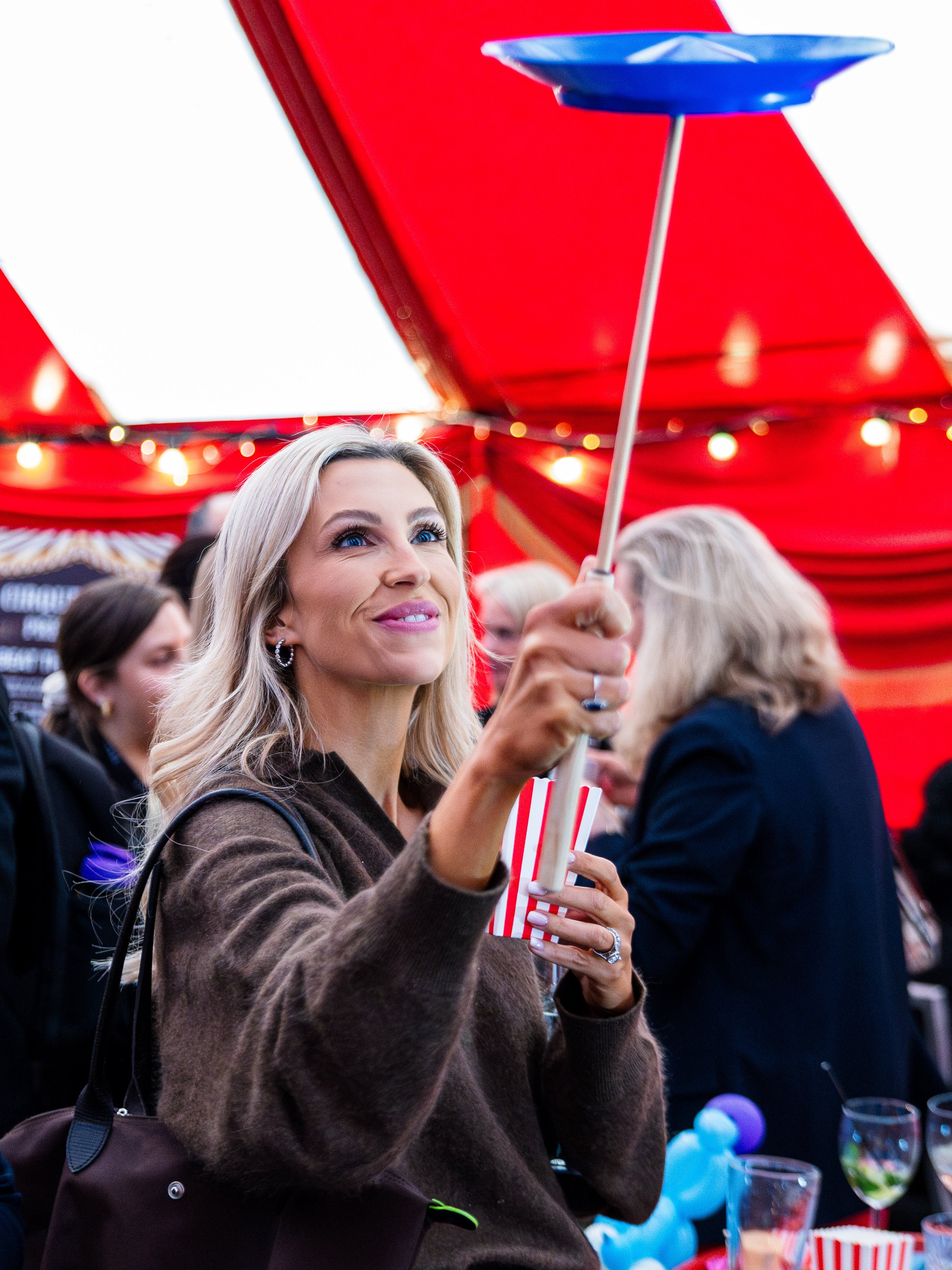 A woman with blonde hair and blue eyes holding a striped straw and an umbrella at an outdoor event with red tents and string lights in the background.