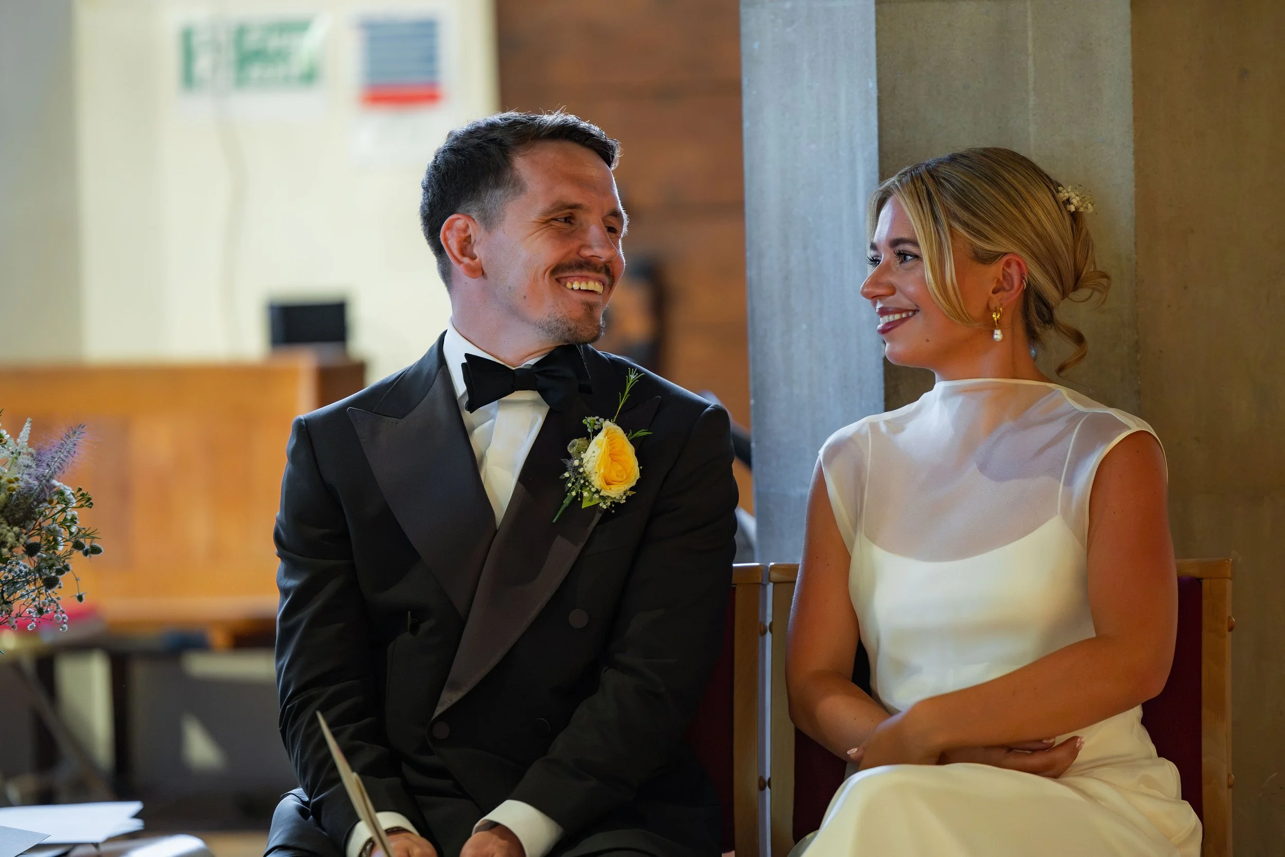 A man dressed in a tuxedo and a woman in a white dress sitting together and smiling at each other.