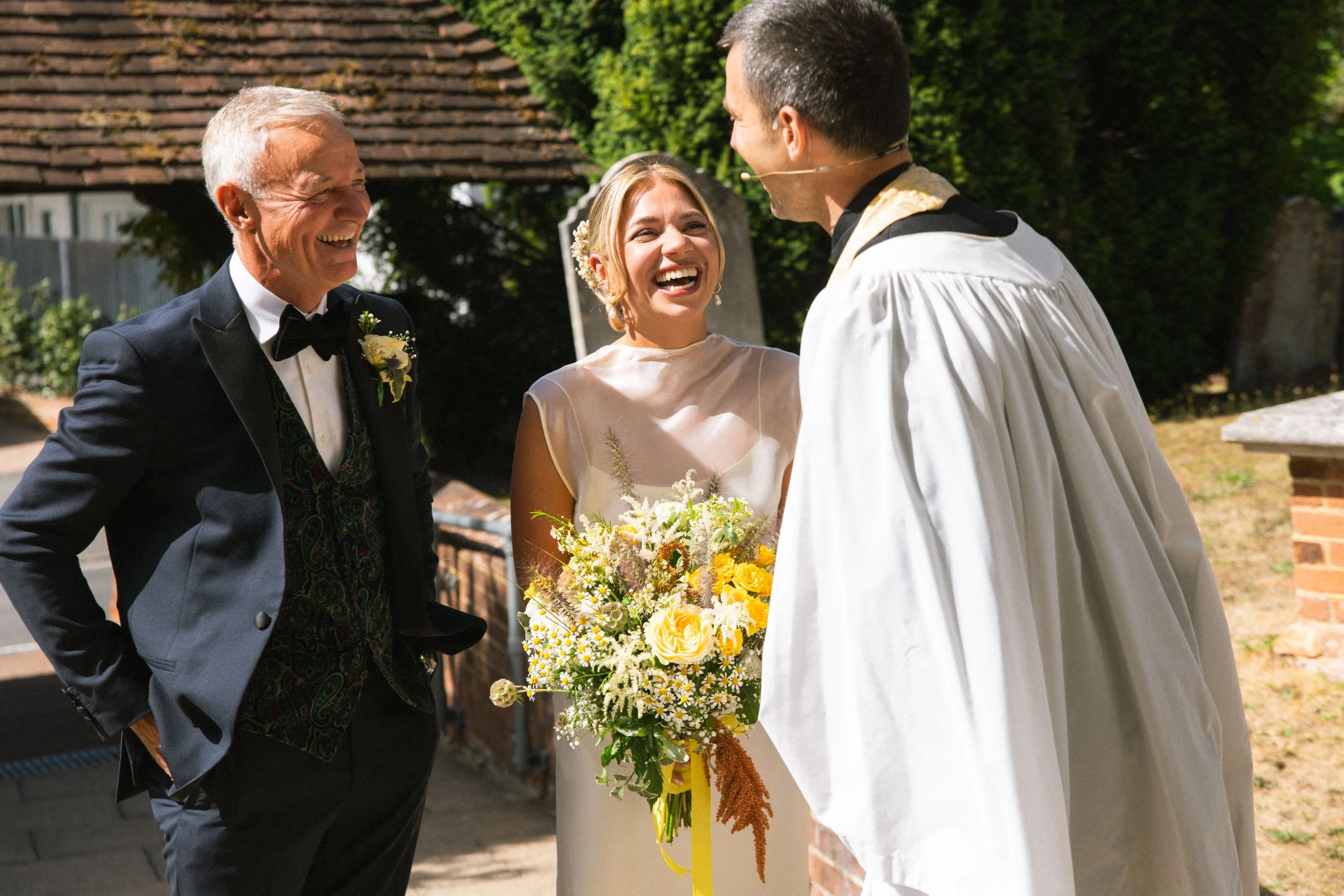 A wedding ceremony with a bride holding a bouquet of yellow and white flowers, smiling at the groom, with an older man in a tuxedo standing nearby and smiling, in an outdoor setting with trees and a brick building in the background.