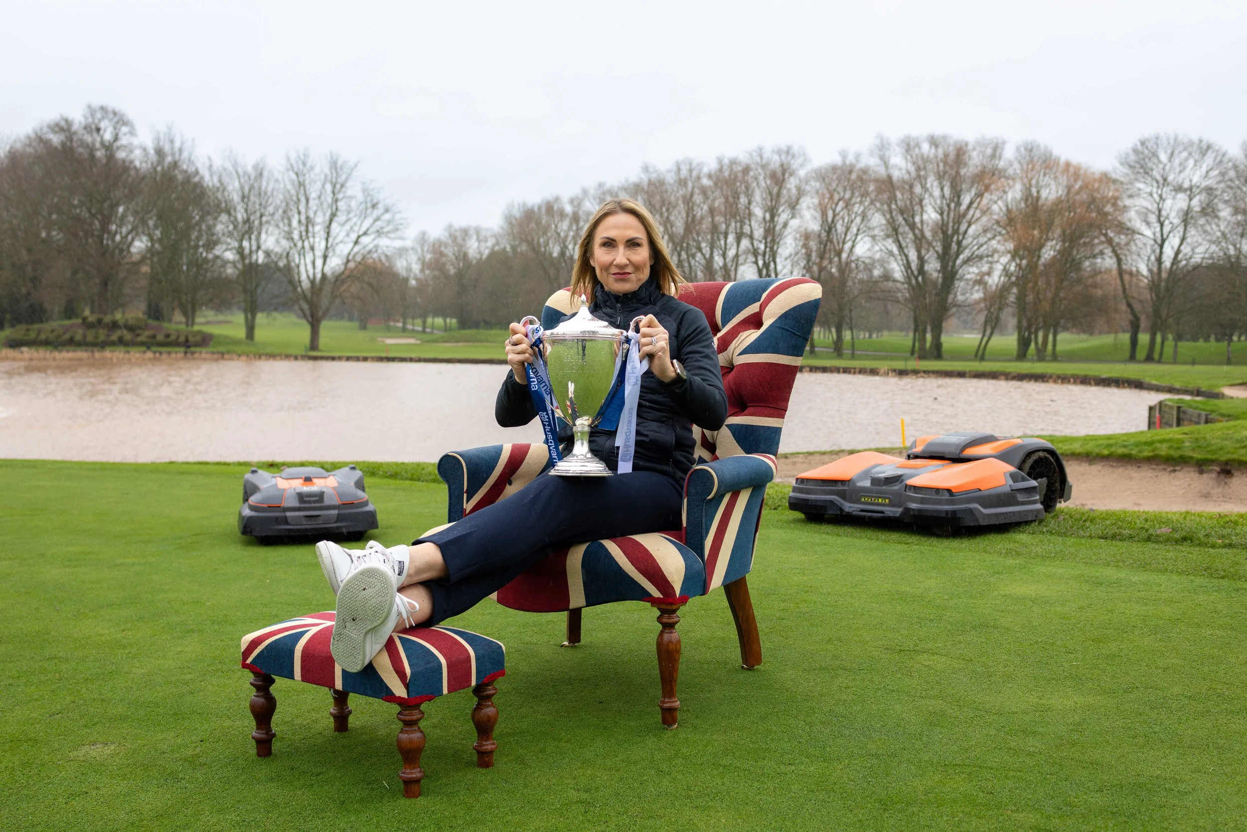 Woman sitting in an armchair outdoors on grass, holding a large silver trophy with sports medals hanging from it. There are robotic lawnmowers around her and a lake with trees in the background.