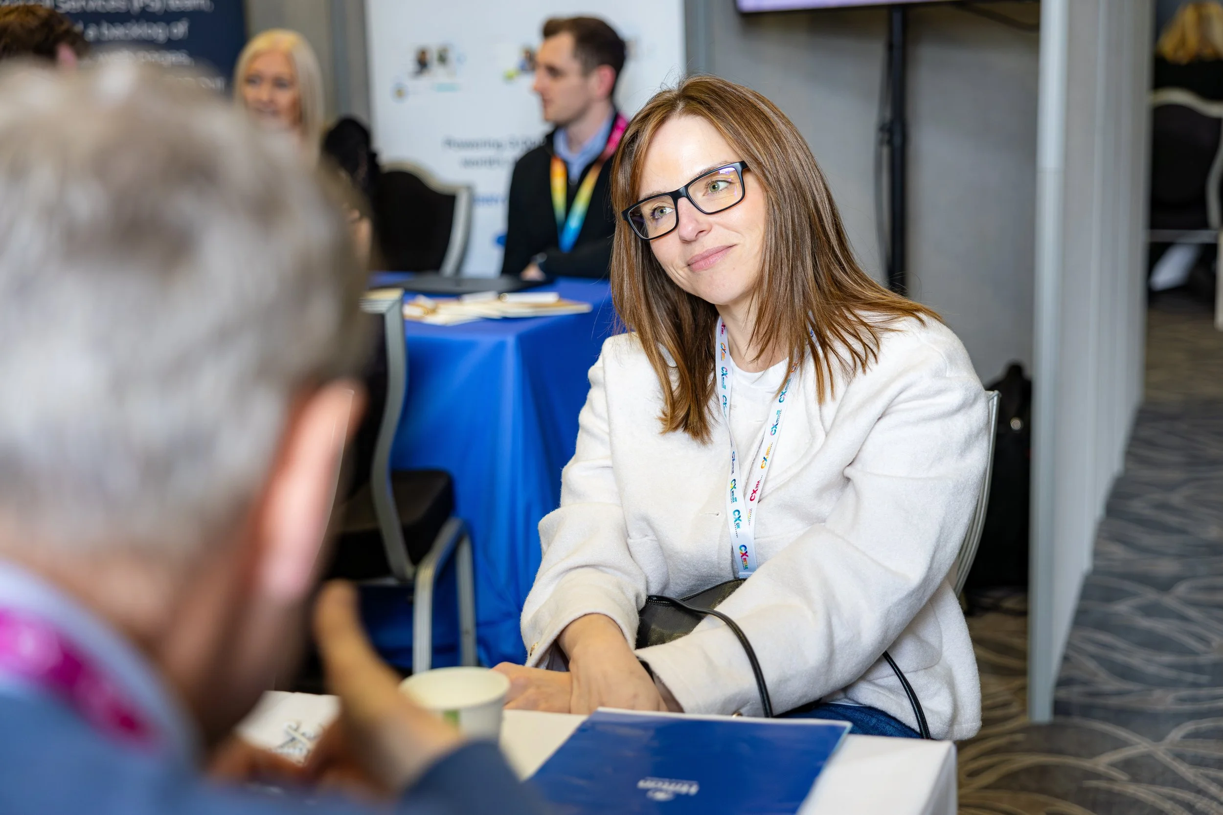 A woman with brown hair, glasses, and a cream-colored sweatshirt sitting at a table, engaging in conversation with someone partially visible in the foreground. She has a lanyard around her neck and appears to be at an event or conference. There are other people and a display board in the background.