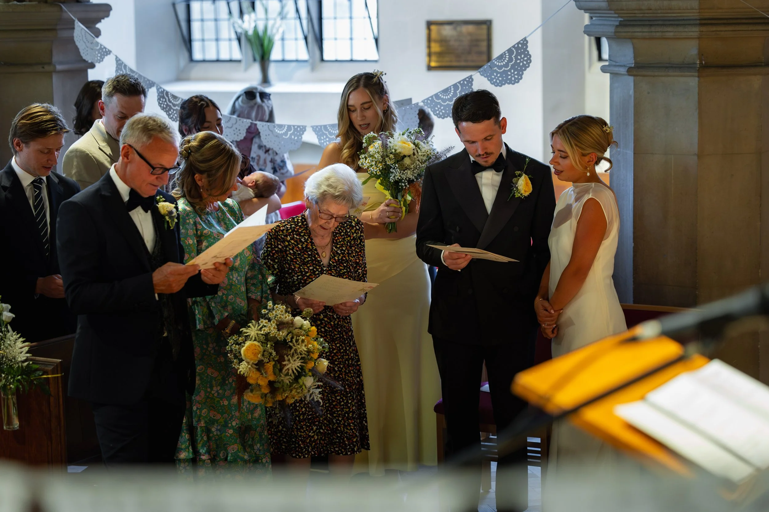 A wedding ceremony with a bride and groom standing before an officiant, surrounded by guests. The bride wears a white dress, and the groom is in a black tuxedo. Guests are holding papers, and one woman is holding a bouquet. Decorations include flower
