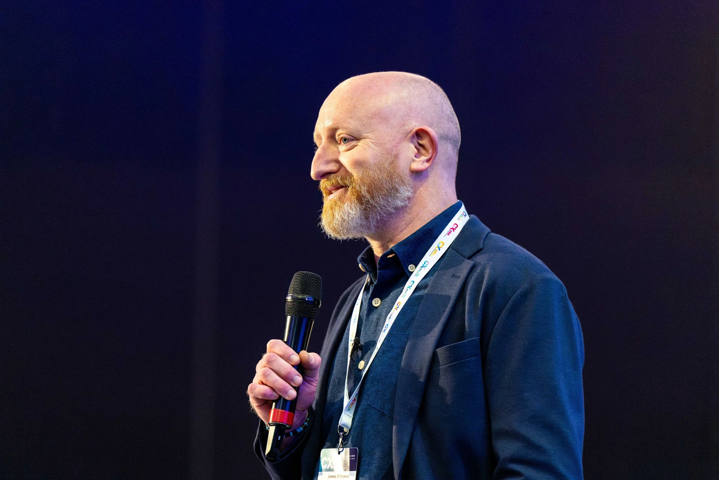 Man with a red beard and shaved head speaking into a microphone during a presentation, wearing a dark jacket and an event badge around his neck.