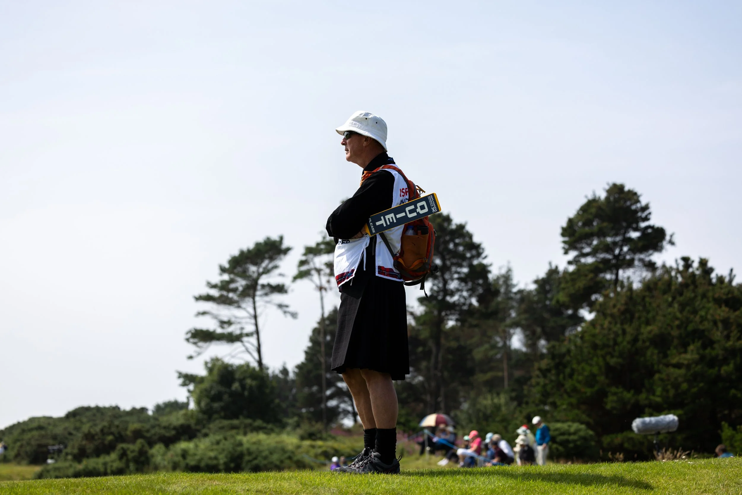 A man wearing a white bucket hat, sunglasses, and a black shirt with a red, white, and blue vest, stands on a golf course holding a golf club. In the background, a group of children and adults are gathered, with trees and a slightly overcast sky over