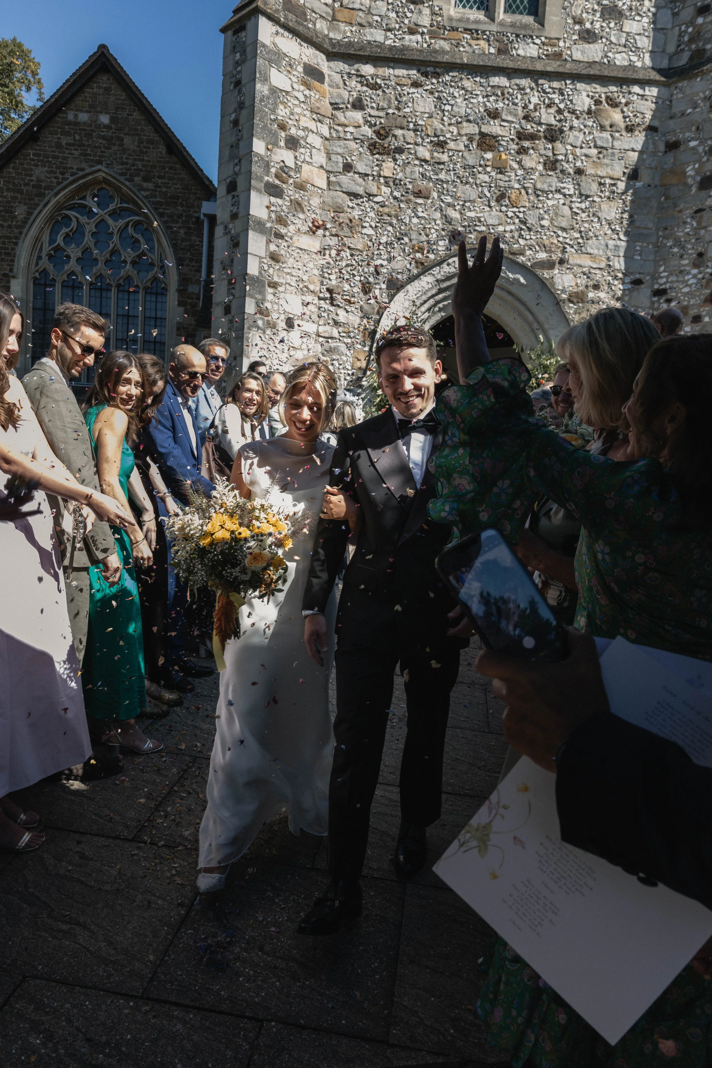 A wedding celebration outside a stone church with a group of guests throwing confetti, including a bride and groom smiling, surrounded by friends and family.