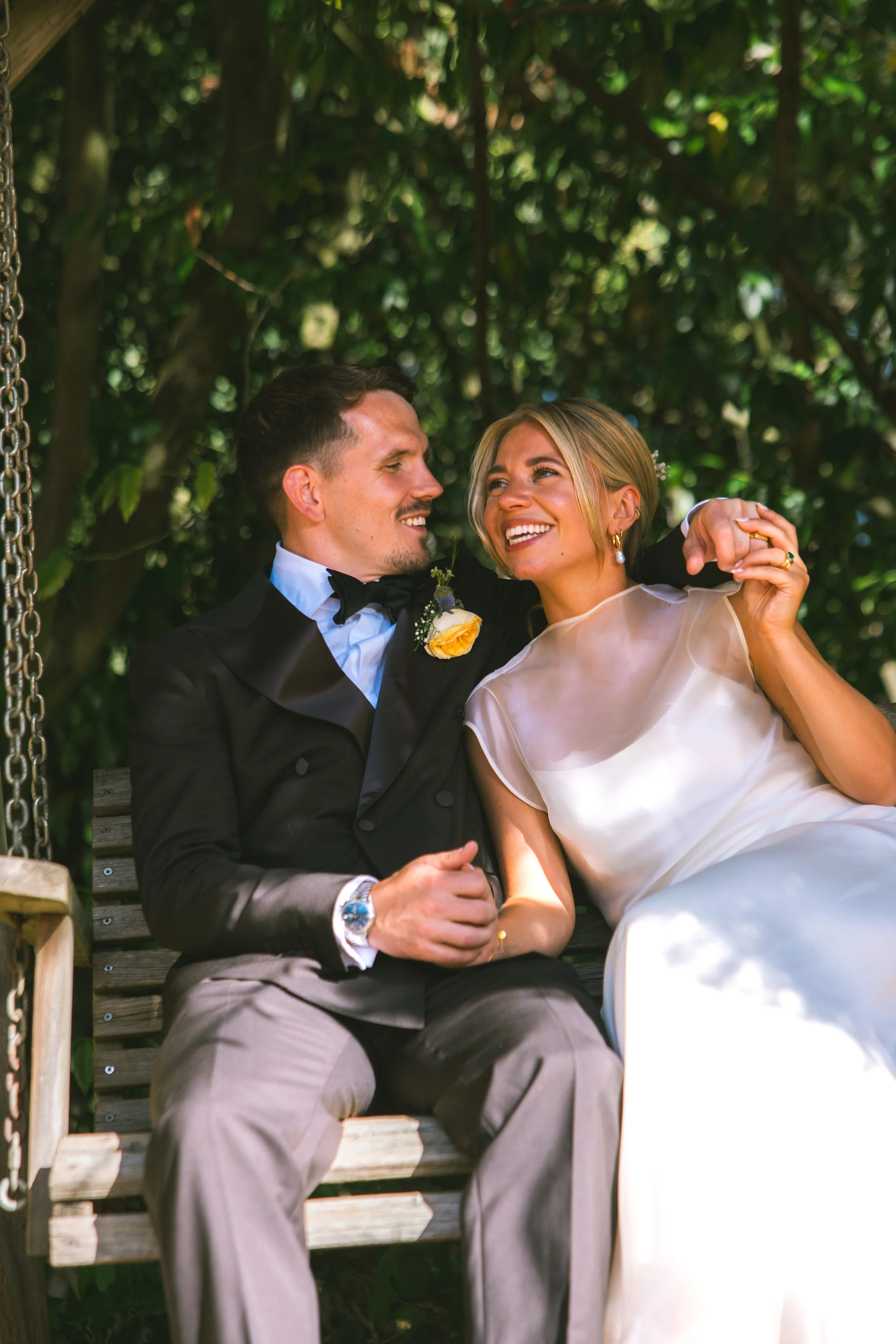 A smiling couple dressed in wedding attire sitting on a wooden bench outdoors surrounded by greenery.