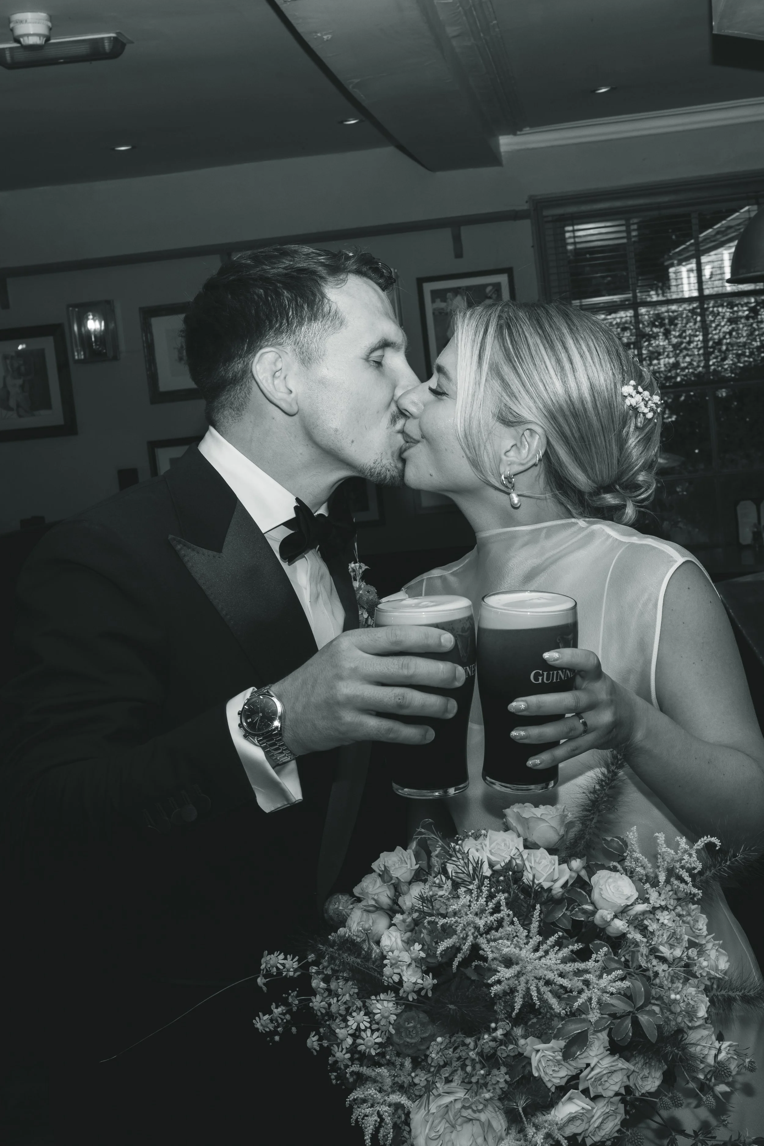 A black and white photo of a newlywed couple sharing a kiss at their wedding reception. They are holding Guinness beer glasses, and there is a floral arrangement on the table in front of them. The background shows framed pictures on the wall and a wi