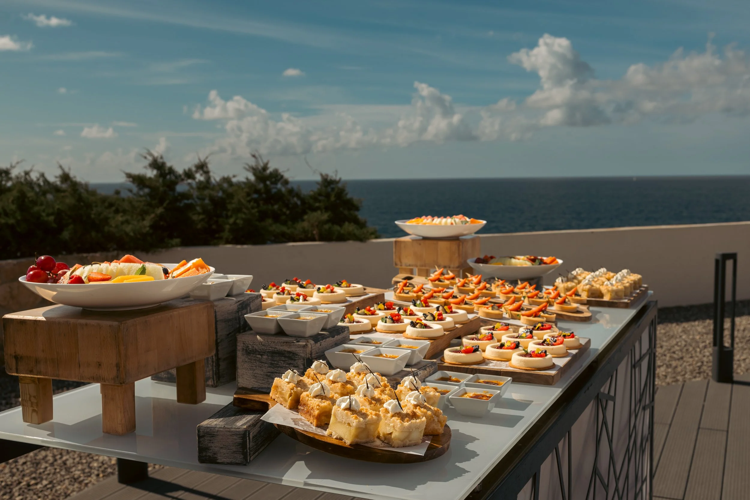 Outdoor table with an assortment of desserts including parfaits, cakes, cookies, and fresh fruit with a scenic ocean view and blue sky in the background.