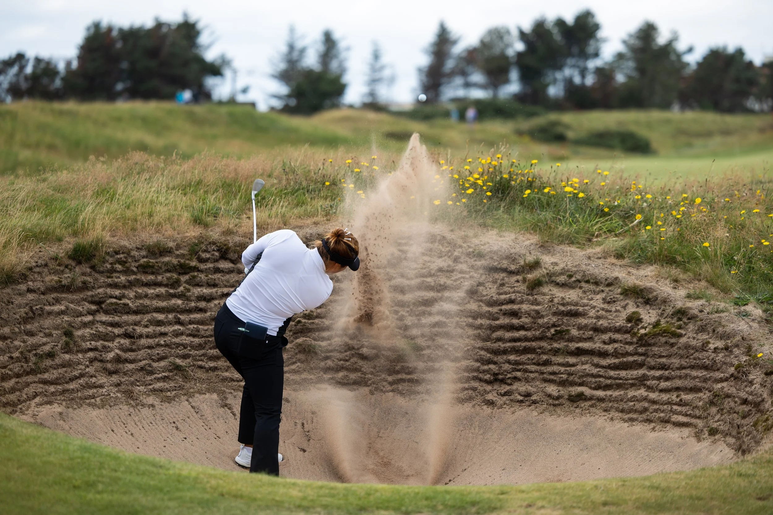 A woman in white golf attire is taking a shot from a sand trap on a golf course, with a sand explosion occurring as she hits the ball.