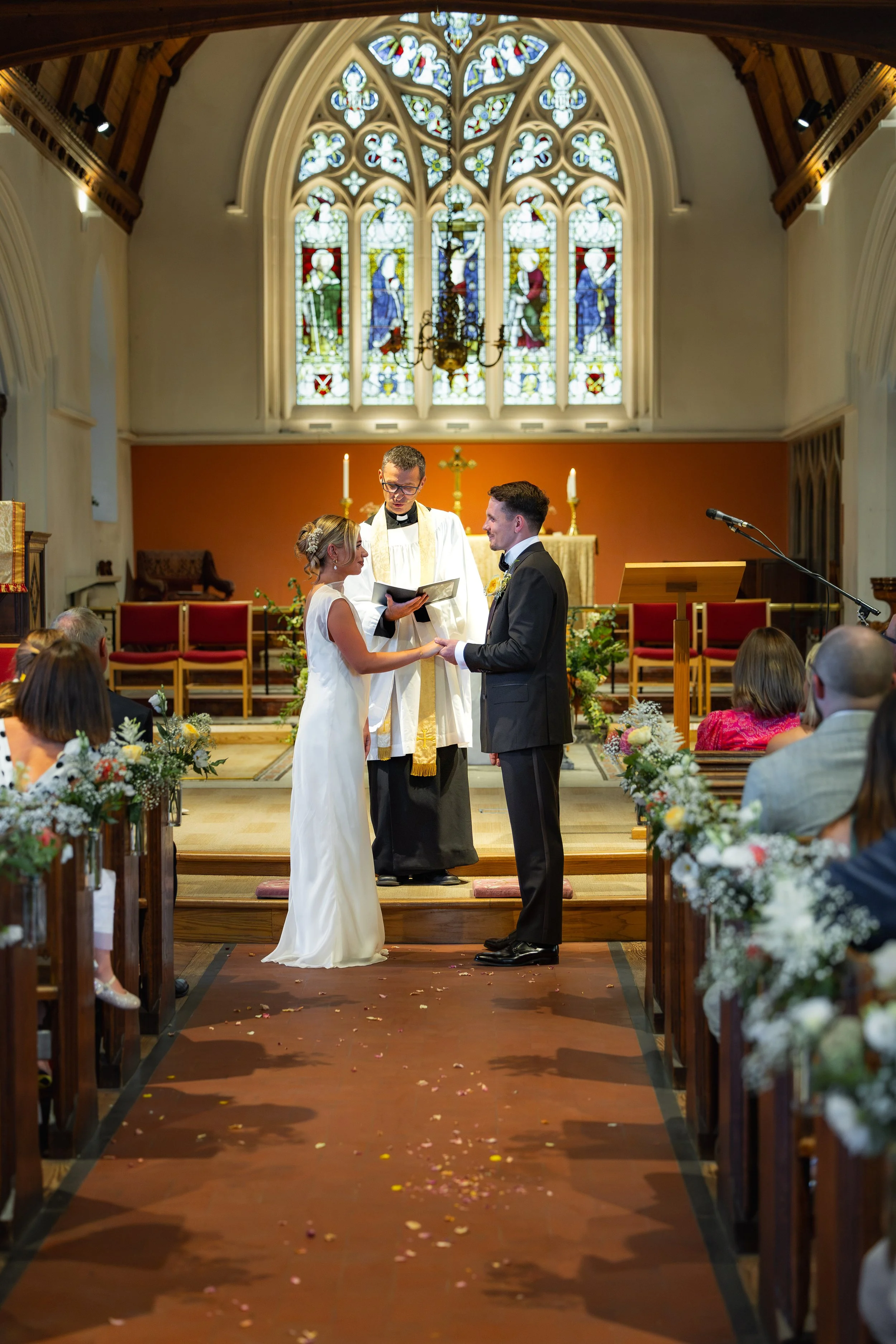 A wedding ceremony inside a church with stained glass windows, a priest officiating, and a couple standing at the altar exchanging vows.